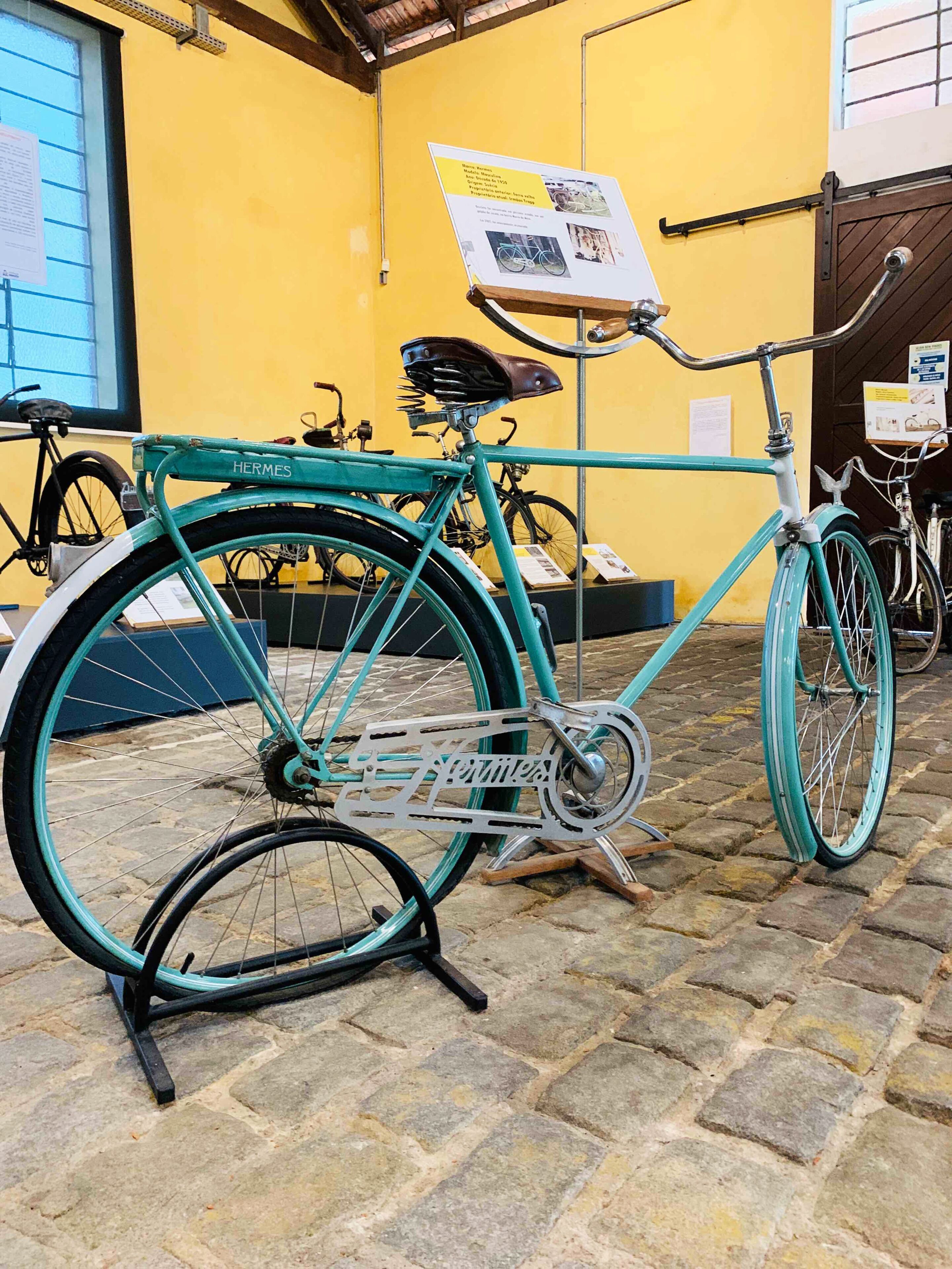 A turquoise Hermes bicycle with a leather saddle and metal frame, displayed indoors against a yellow wall with informational posters.