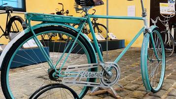 Una bicicleta turquesa de Hermès con sillín de cuero y estructura metálica, exhibida en un interior frente a una pared amarilla con carteles informativos.