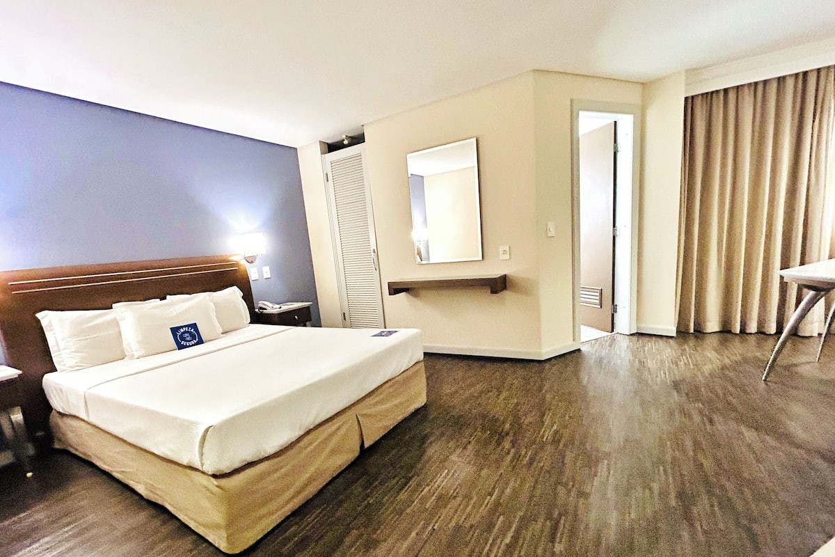A hotel room with a large bed, a desk, a chair, a mirror, and a door leading to another room.