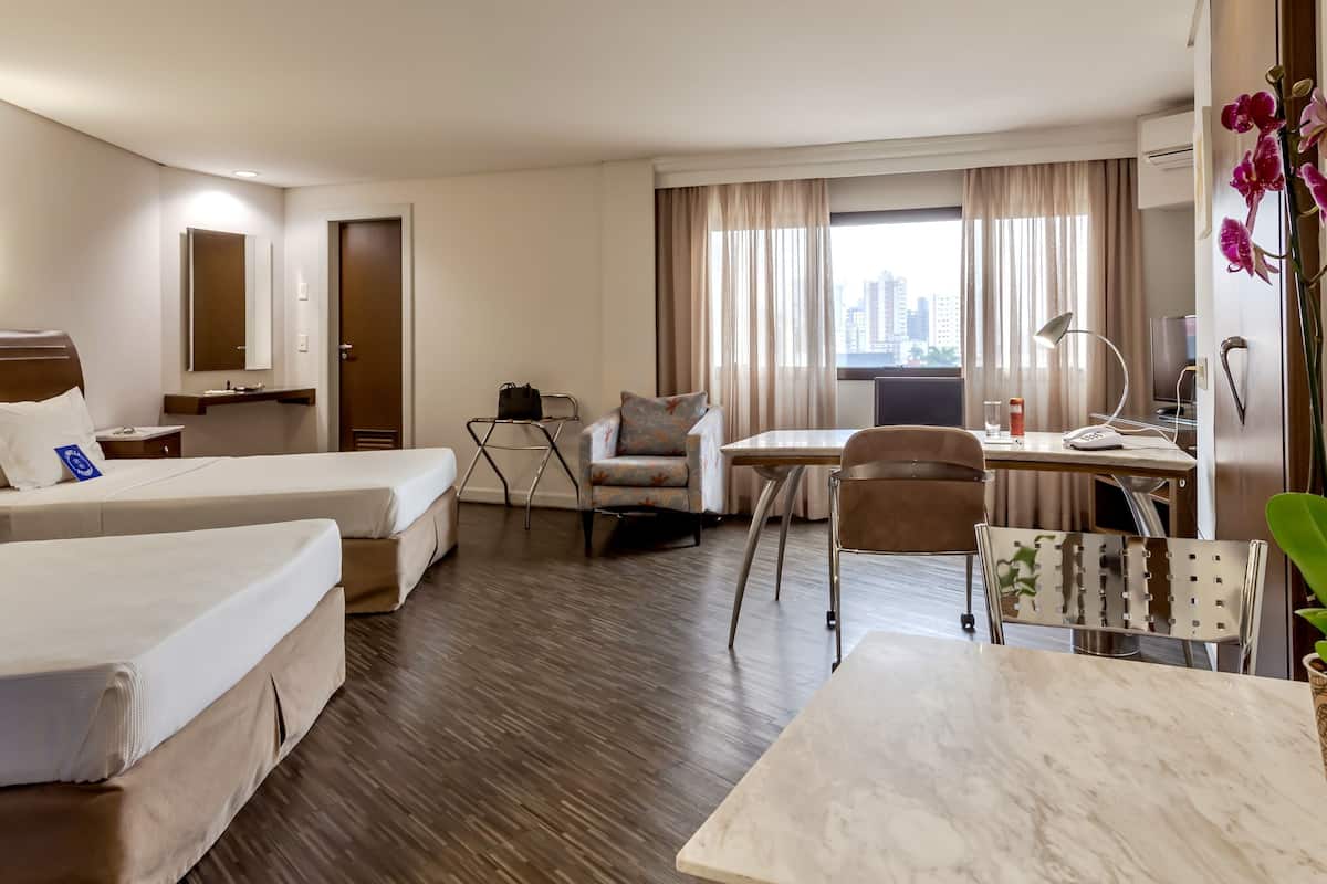 A hotel room with two beds, a desk, a chair, a small table, a lamp, and a view of the city.