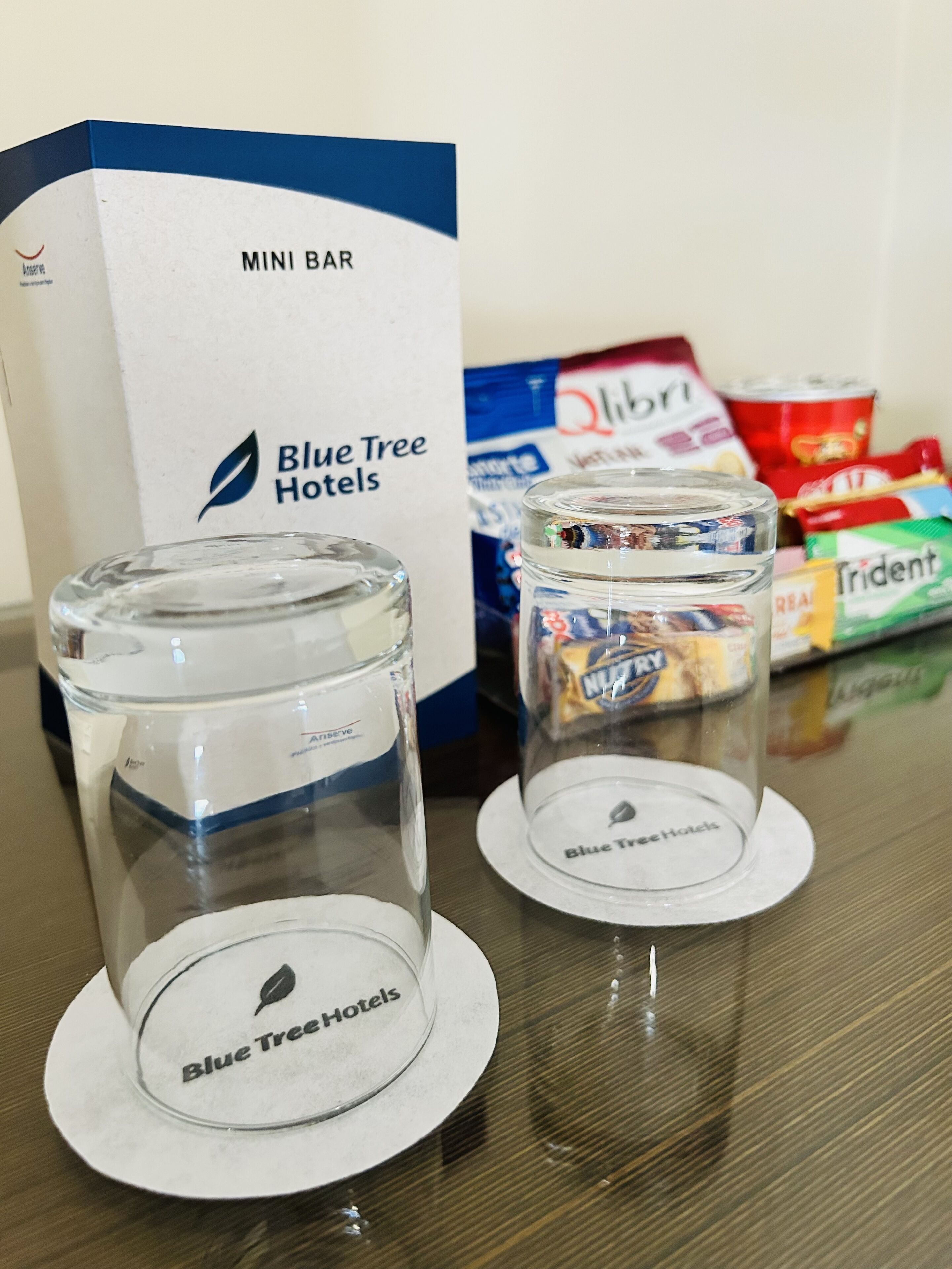 Two clear glasses on coasters with the Blue Tree Hotels logo, a mini bar box, and snack packages in the background.
