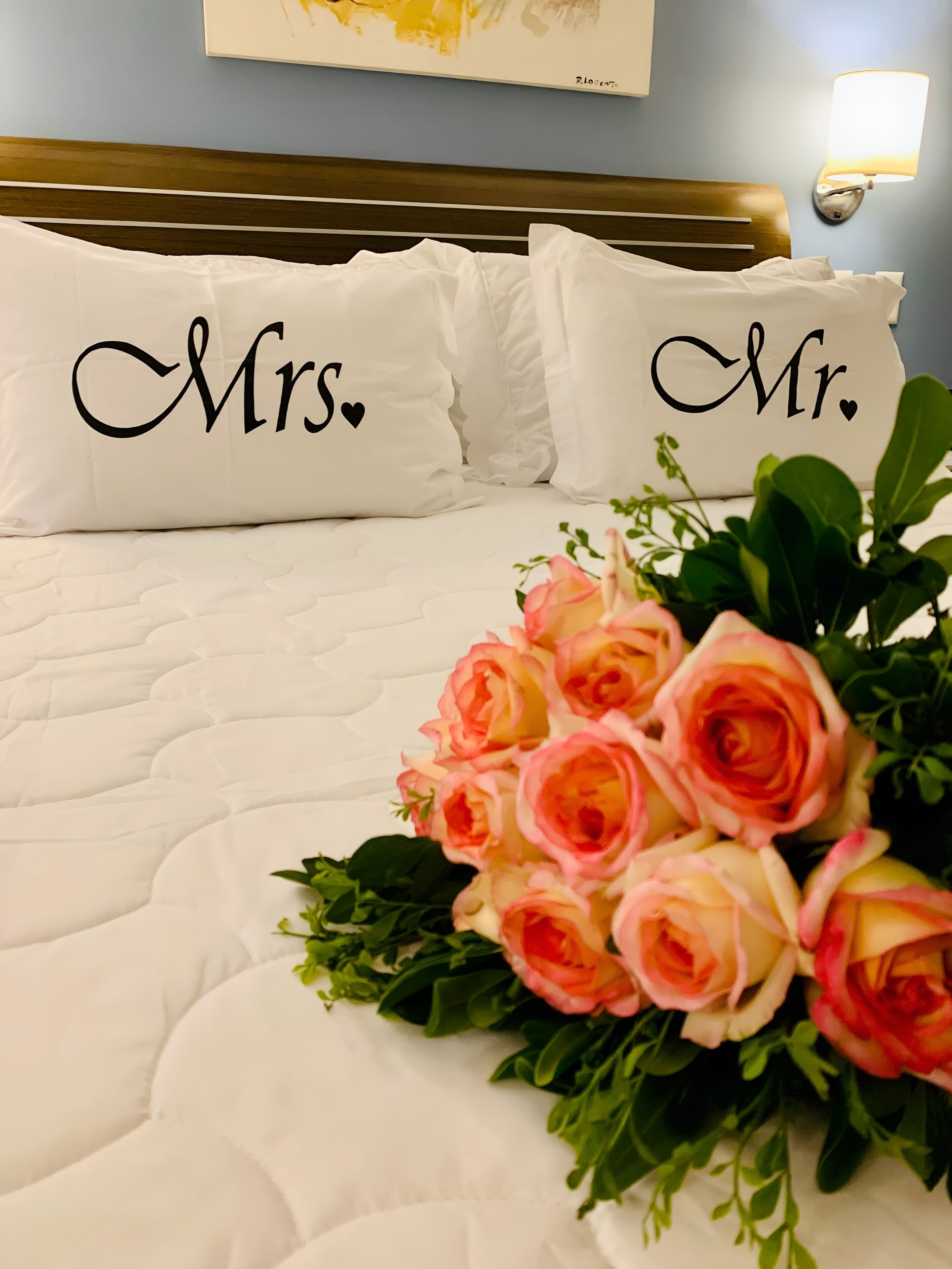 Uma cama com almofadas rotuladas "Mrs." e "Mr.", um buquê de rosas cor-de-rosa e uma luminária na parede.