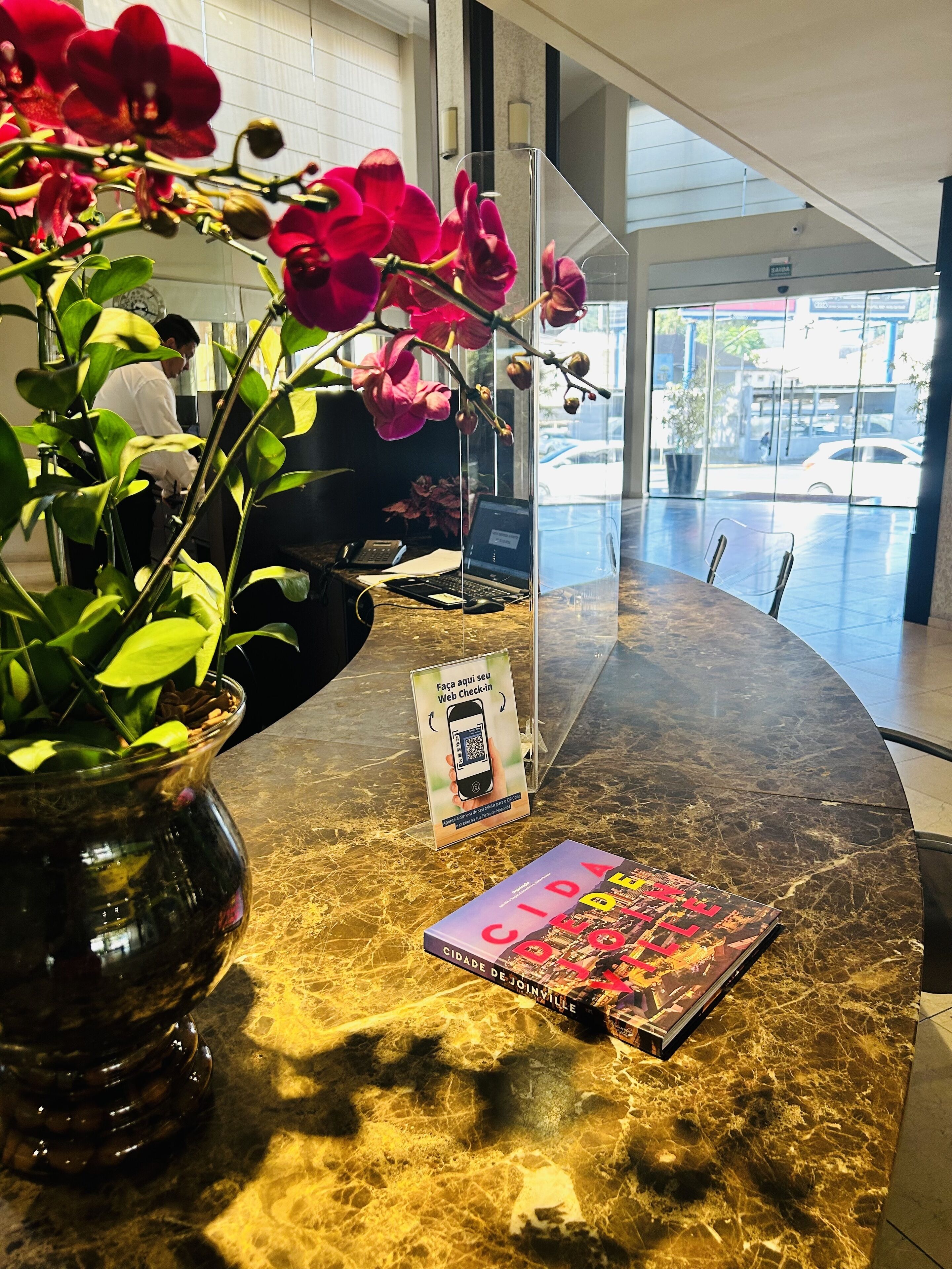 A marble reception desk with a laptop, a QR code sign, a book titled 'CIDADE DE JOINTVILLE', and a vase with red flowers.