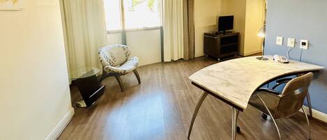 A hotel room with a wooden floor, a curved marble table, a single chair, a television on a stand, and a window with curtains.