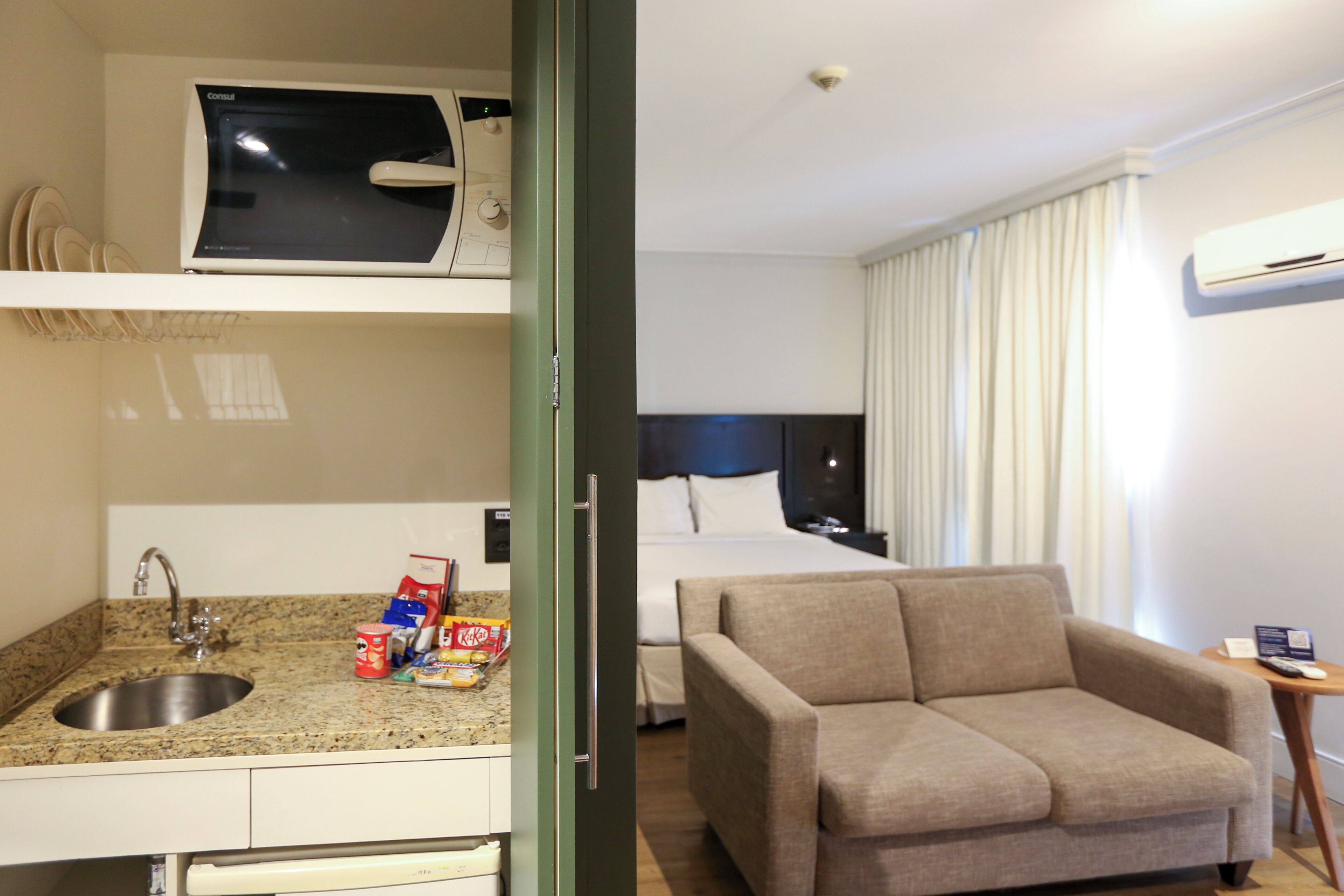 Een hotelkamer met een kitchenette, een bed, een slaapbank en een raam met gordijnen.