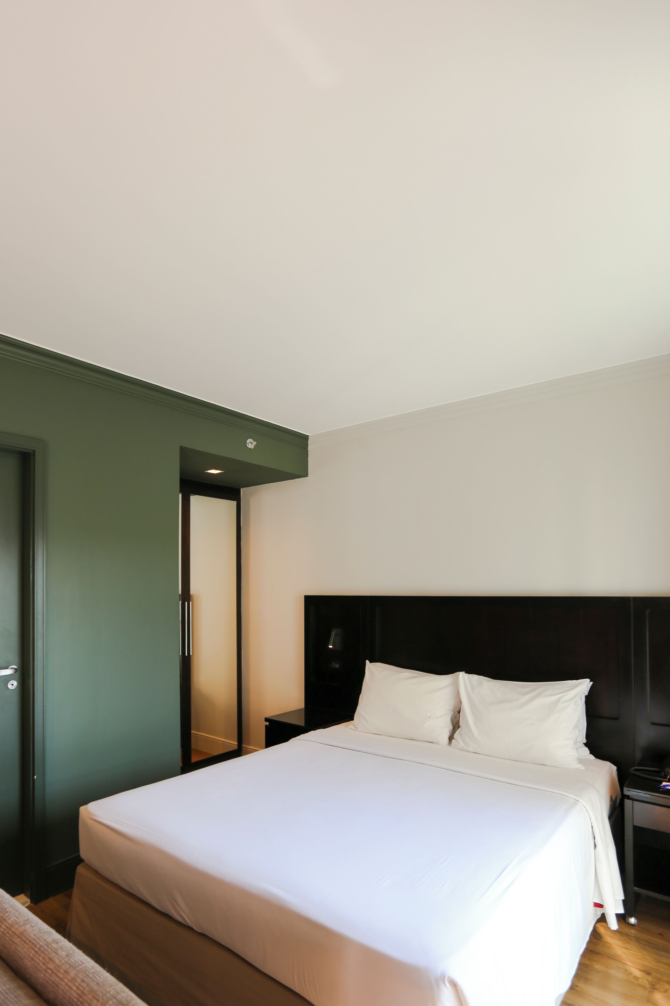 A modern hotel room with a large bed, a dark headboard, and a green accent wall.