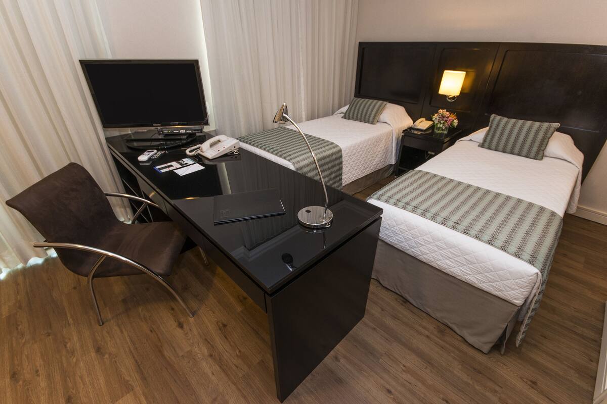 A hotel room with a desk, two beds, a TV, a phone, and a nightstand with a lamp.
