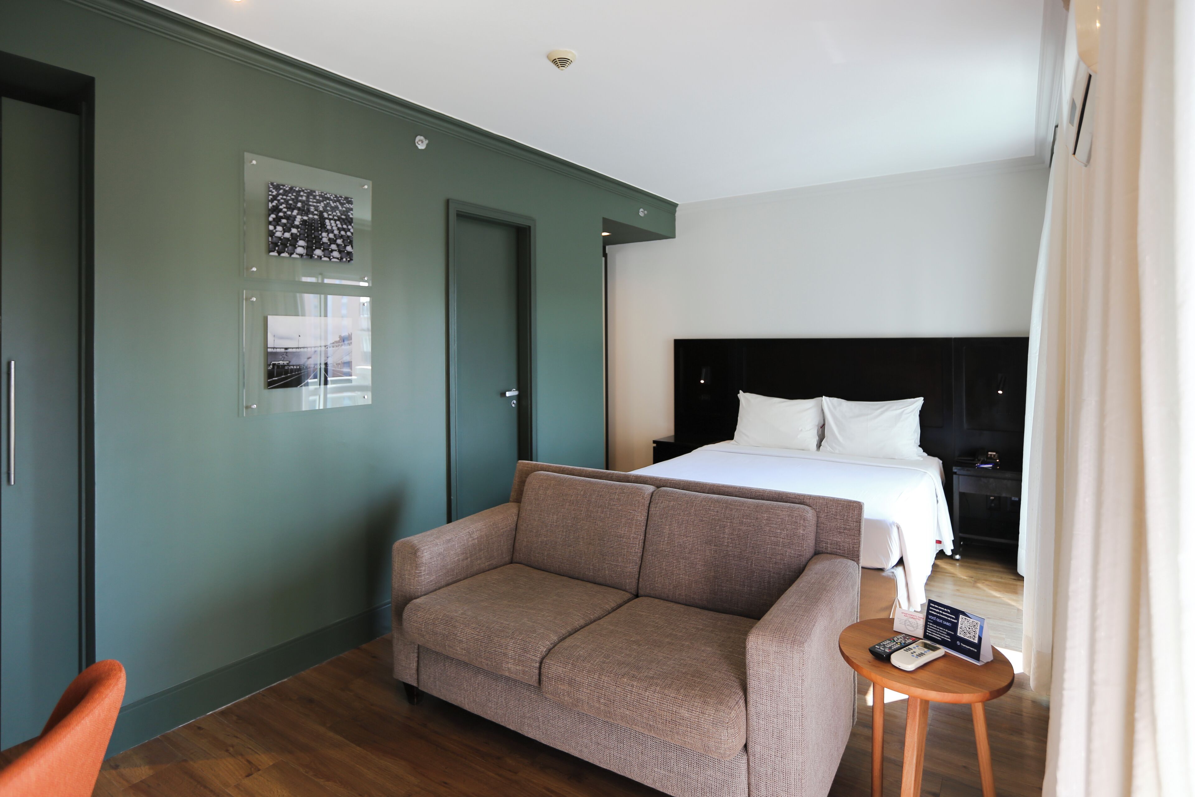 A hotel room with a bed, a sofa, a small table with a remote control, and a door.