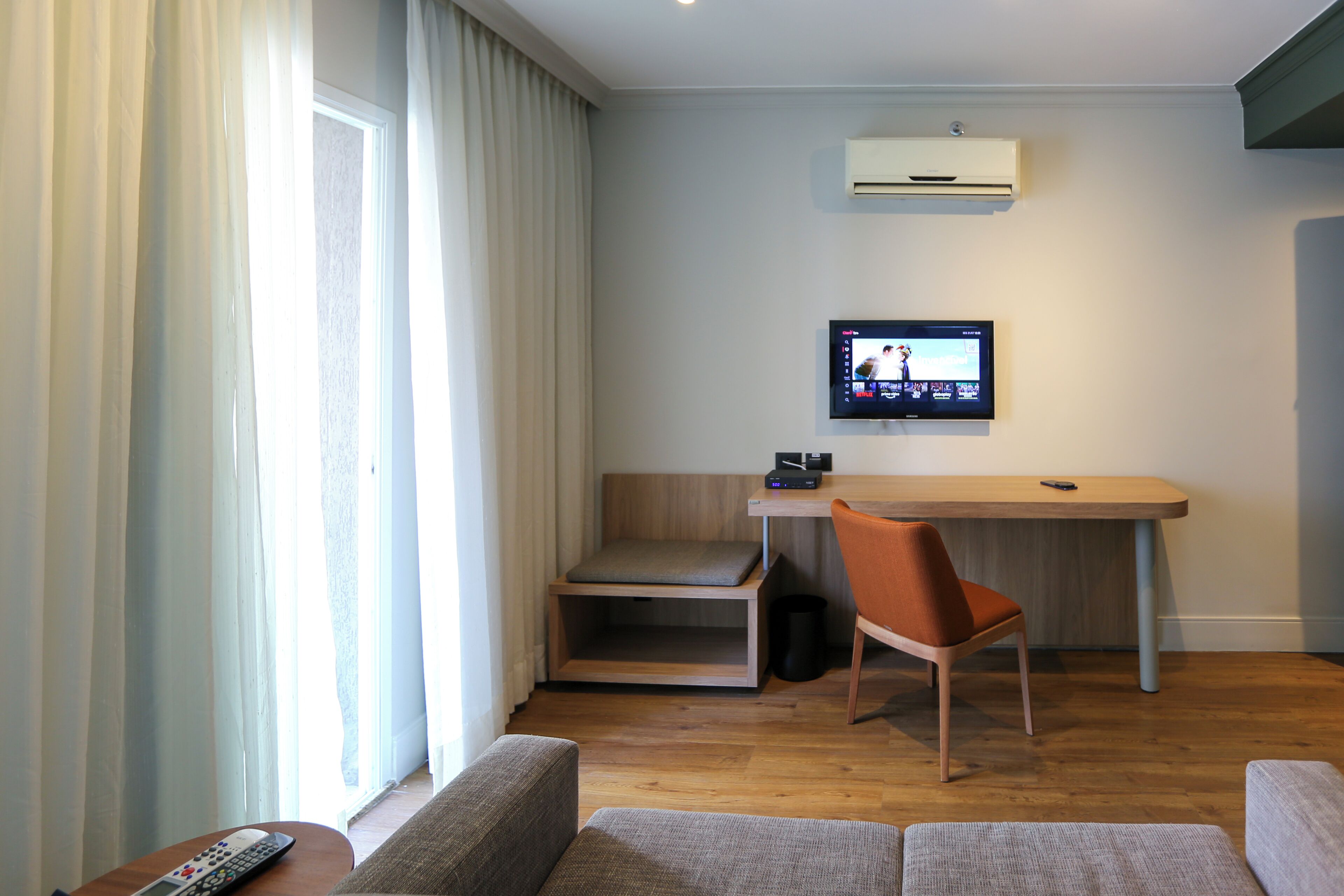 A modern hotel room with a wooden desk, a comfortable chair, a sofa, and a TV mounted on the wall.