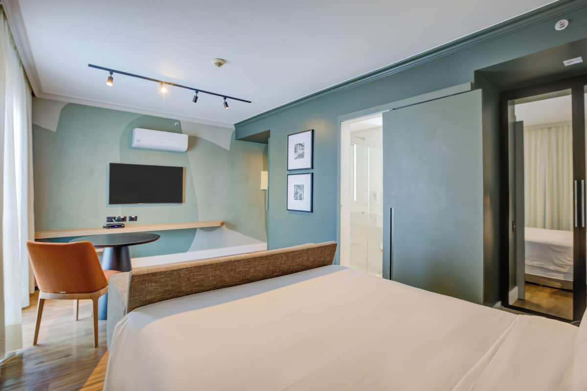 A hotel room with a bed, a desk, a chair, a TV, and a door leading to a bathroom.