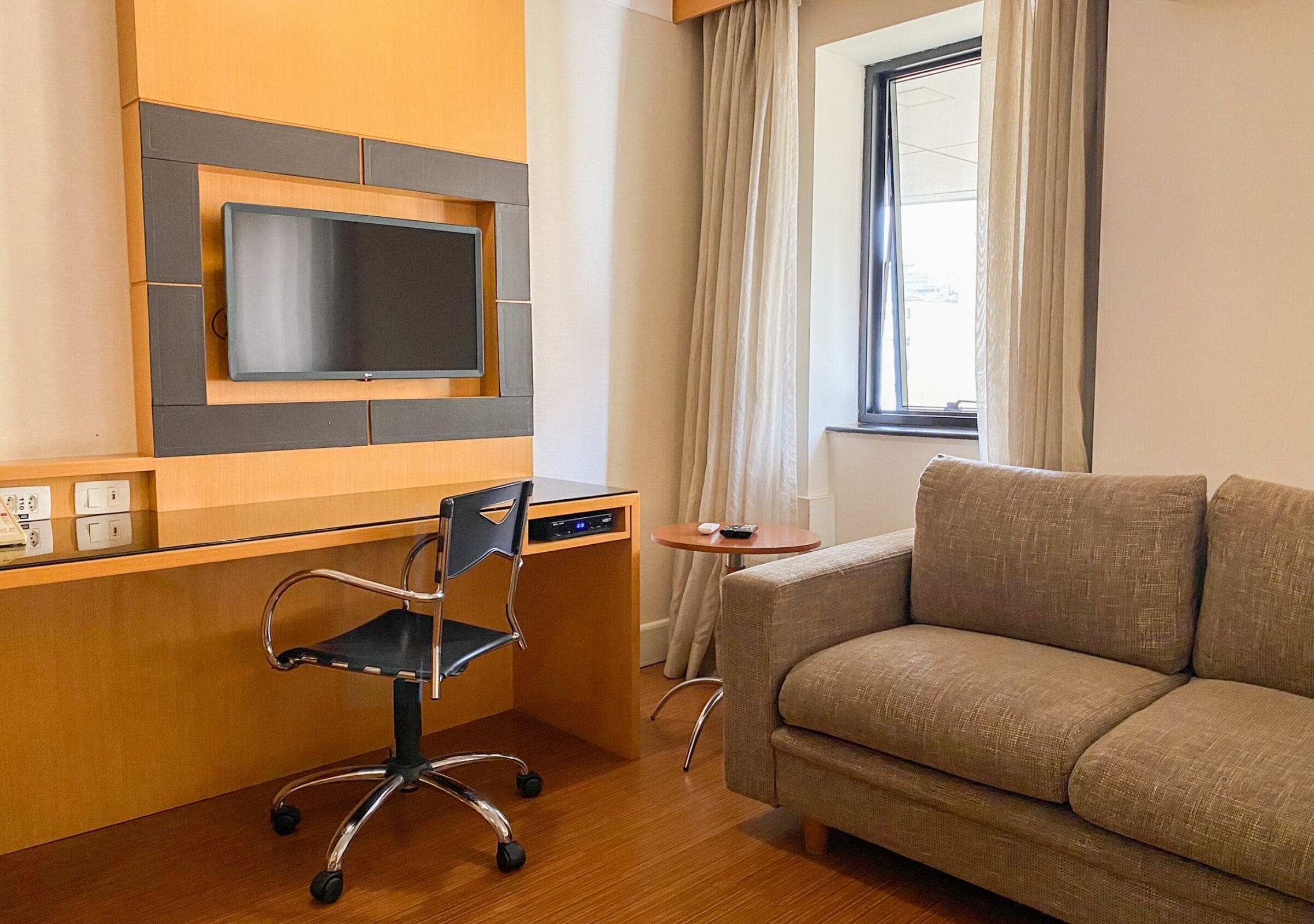 Quarto de hotel com escrivaninha de madeira, cadeira de escritório preta, sofá bege, televisão na parede e janela com cortinas.