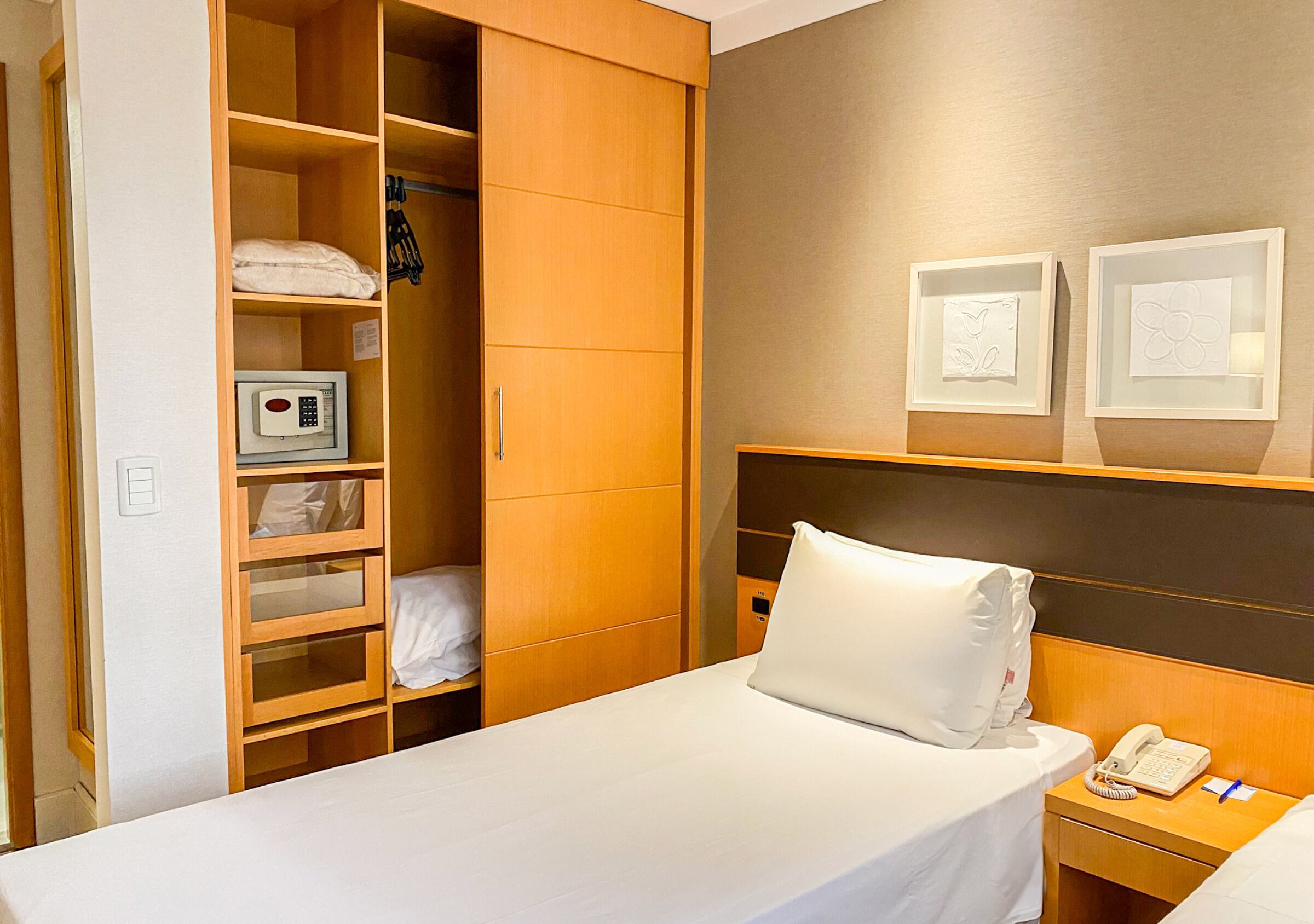 A hotel room with a bed, a bedside table with a phone and pen, a wardrobe with shelves, and a microwave.