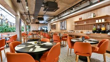 Un restaurante moderno con mesas redondas y sillas naranja, una zona de bar con estantes llenos de botellas, y un amplio ventanal con vistas a la vegetación.