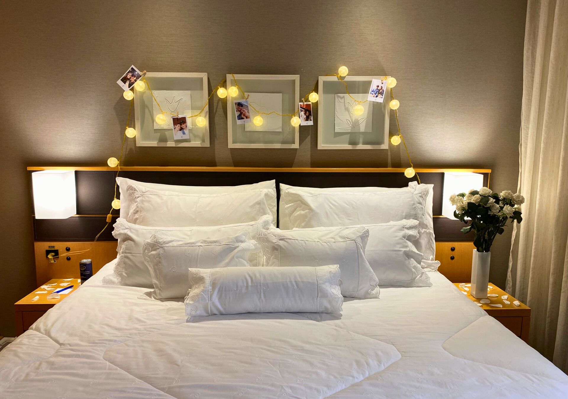 Cama bem arrumada com lençóis brancos, mesa de cabeceira com abajur e parede com fotos emolduradas e luzes de cordão.