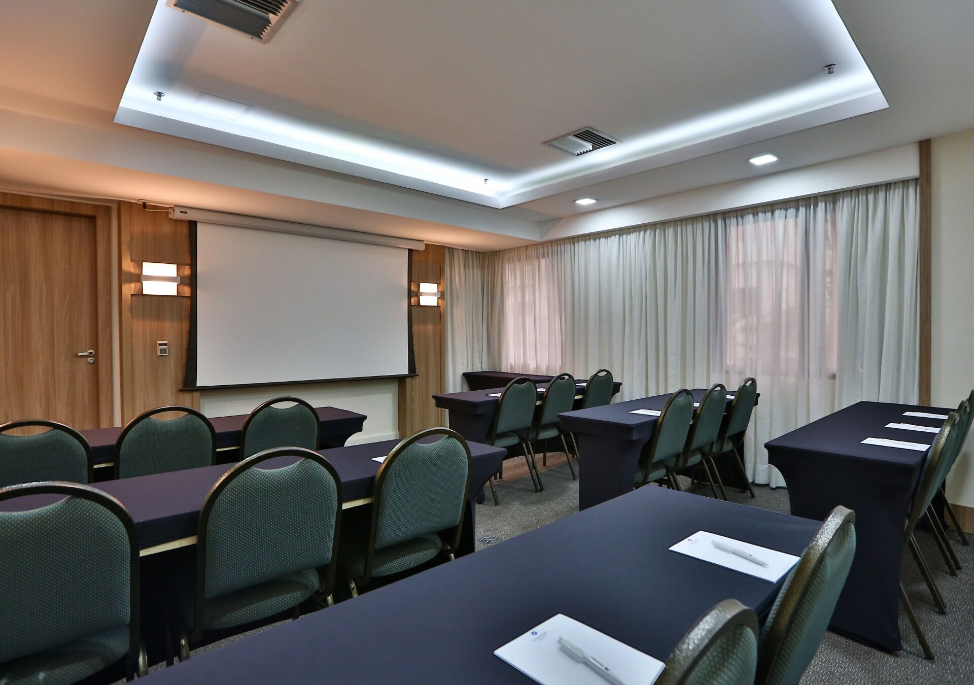 Un salón de conferencias con pantalla de proyector, mesas, sillas y un amplio ventanal con cortinas.