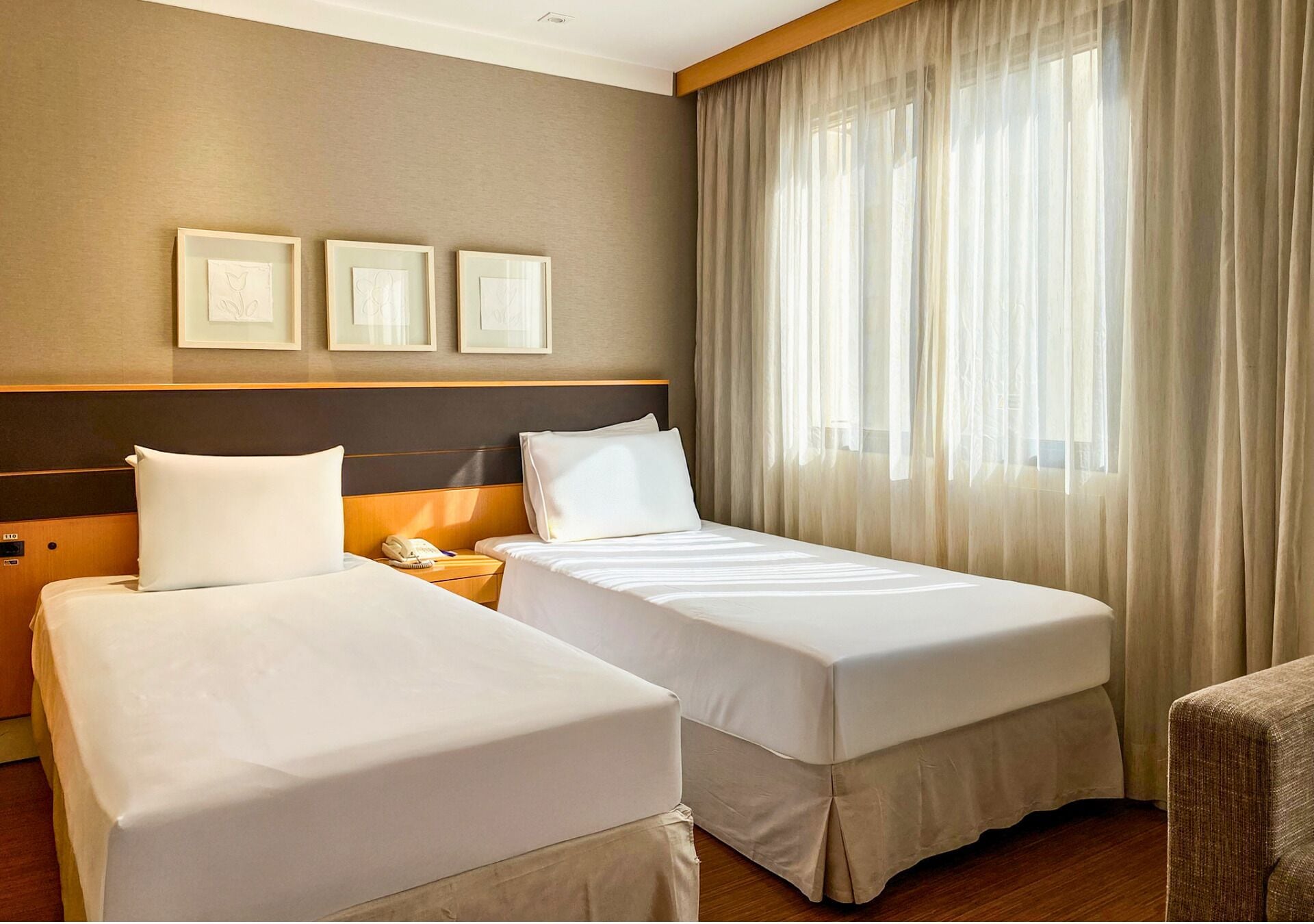 Une chambre d’hôtel avec deux lits simples, une tête de lit en bois, une table de chevet avec un téléphone et une grande fenêtre avec des rideaux légers.
