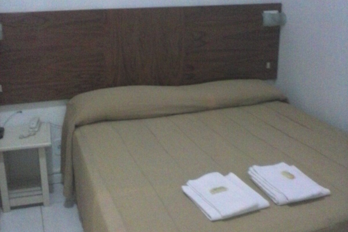Habitación de hotel con cabecera de madera, cama con colcha color crema, mesita de noche con lámpara y dos toallas dobladas sobre la cama.