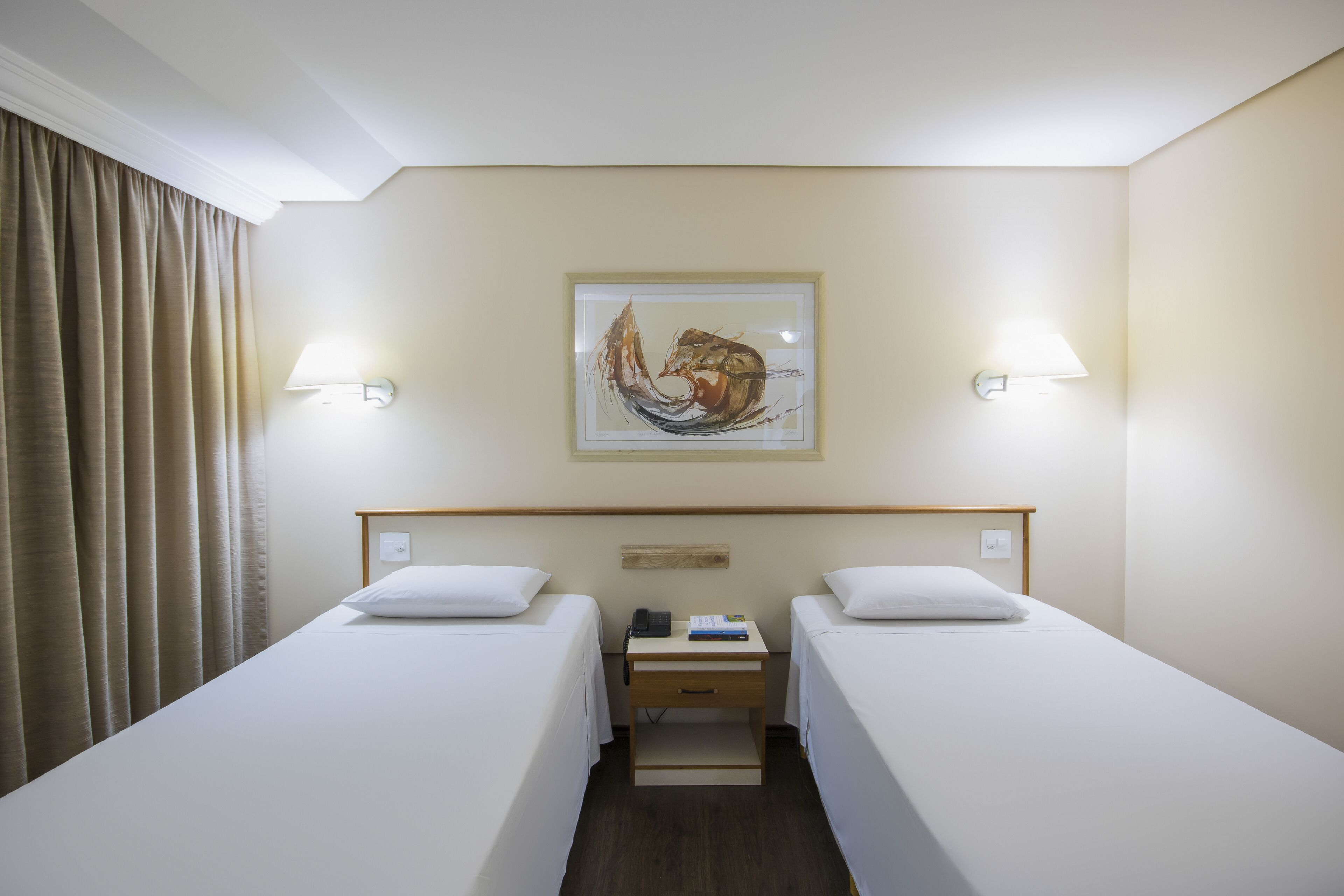 Une chambre d’hôtel avec deux lits simples, une table de chevet avec une lampe, un tableau au mur et une fenêtre avec des rideaux.
