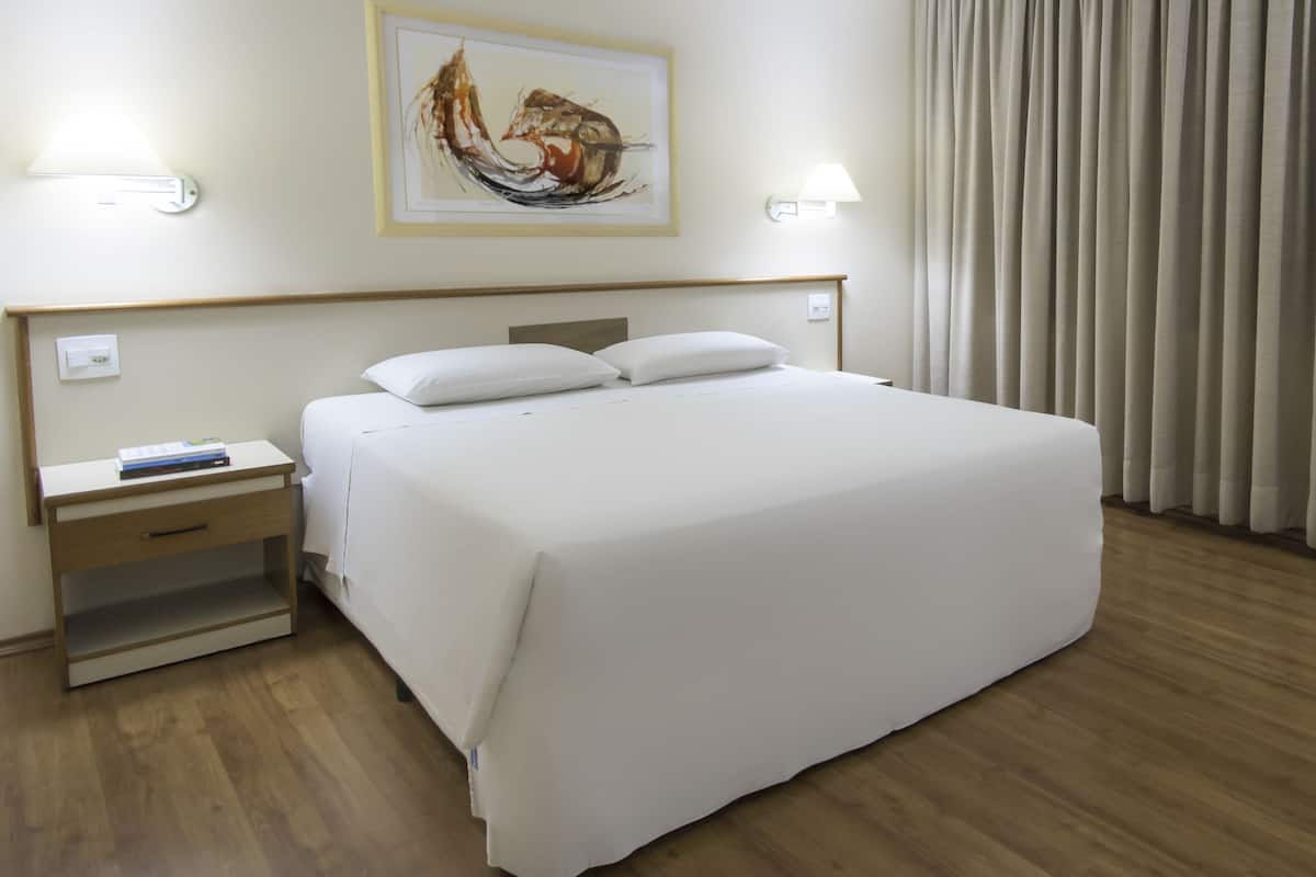A neatly made bed with white linens, a bedside table with a book, and a wall-mounted lamp.