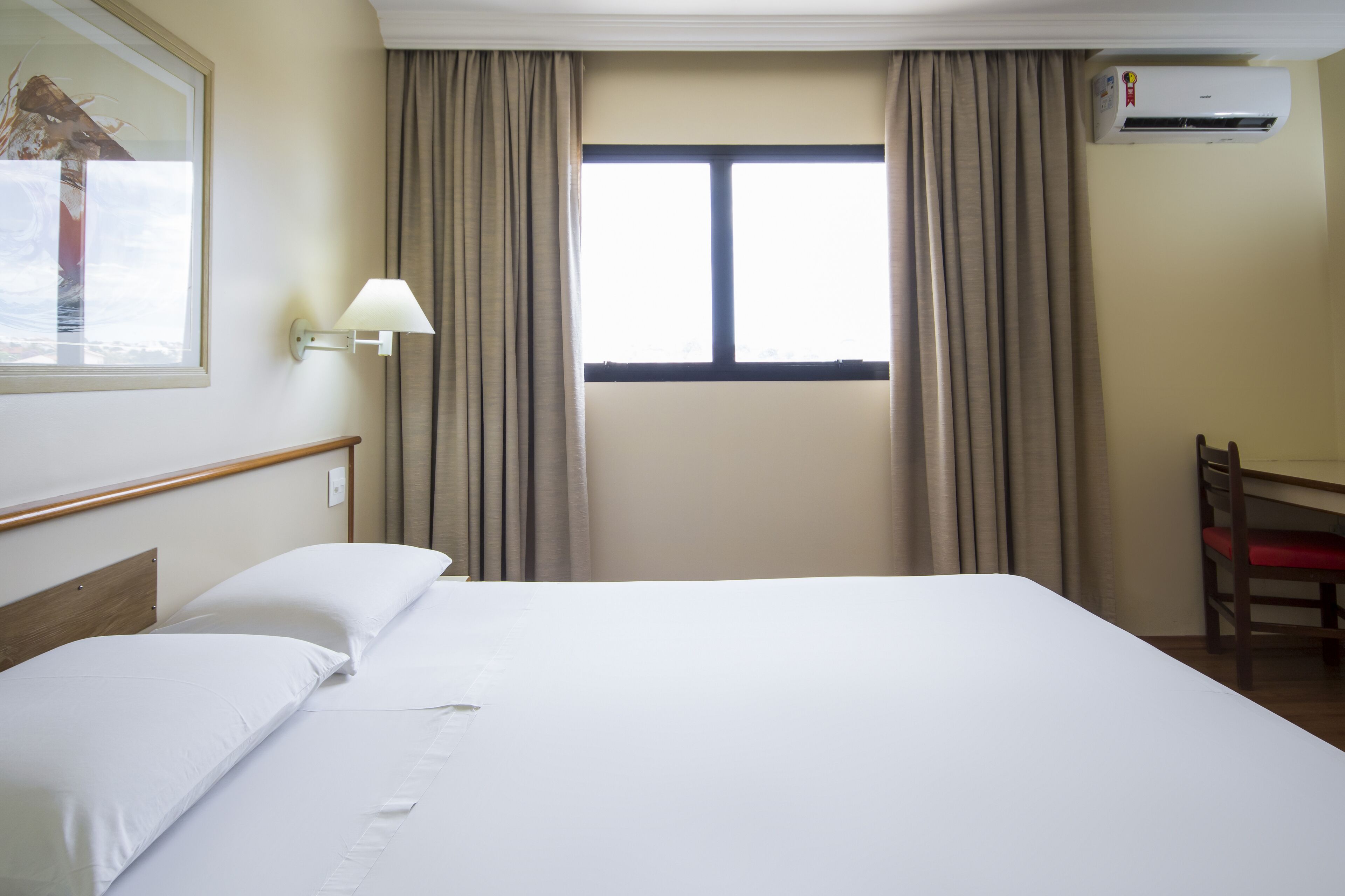 A hotel room with a bed, a desk, a chair, a window with curtains, and an air conditioning unit.
