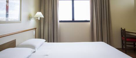 A hotel room with a bed, a desk, a chair, a window with curtains, and an air conditioning unit.