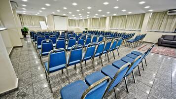 Uma sala de conferências com fileiras de cadeiras azuis, uma tela de projetor e um sofá cinzento.