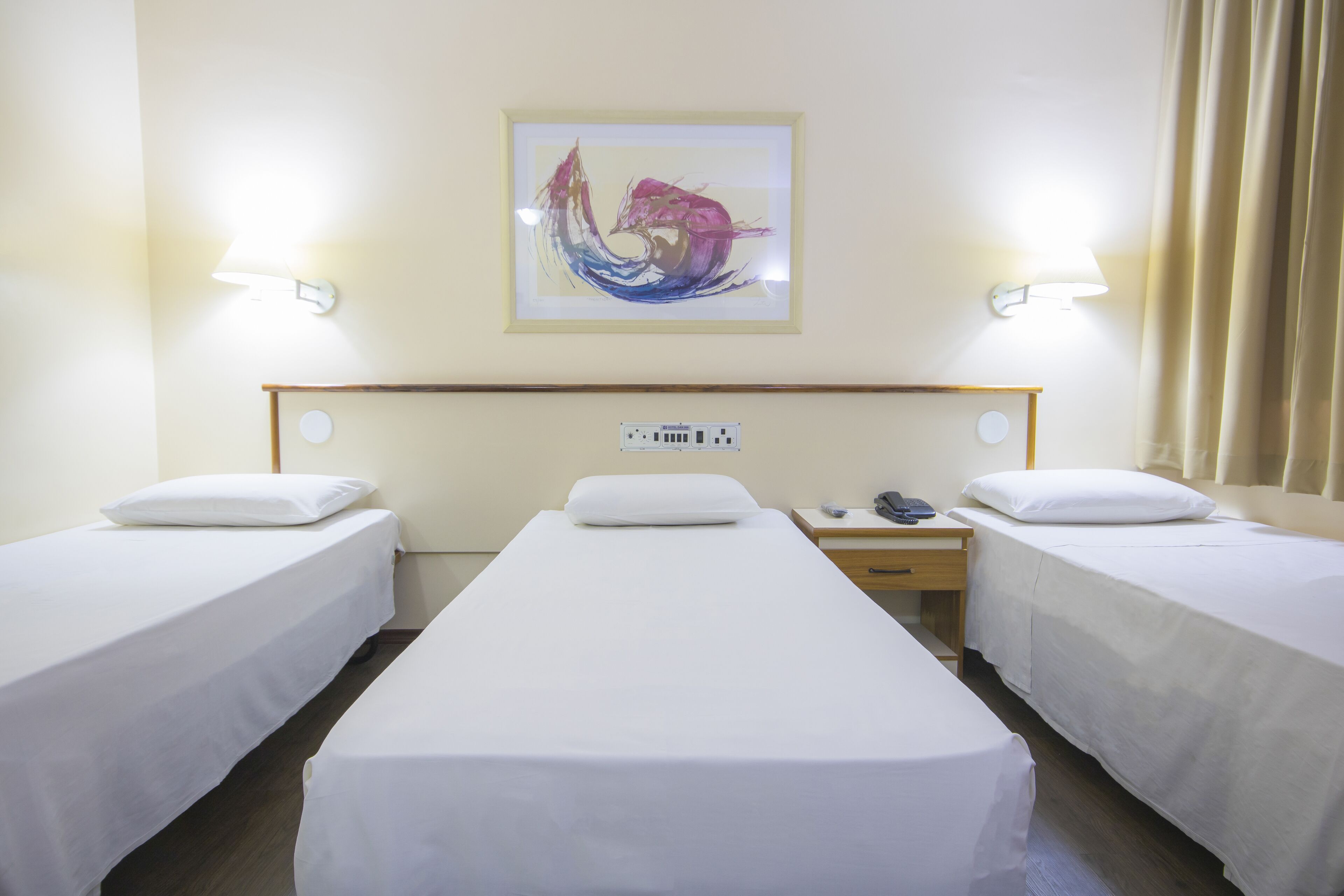 Camera d'albergo con due letti singoli, un comodino con lampada, telefono e presa elettrica a muro.
