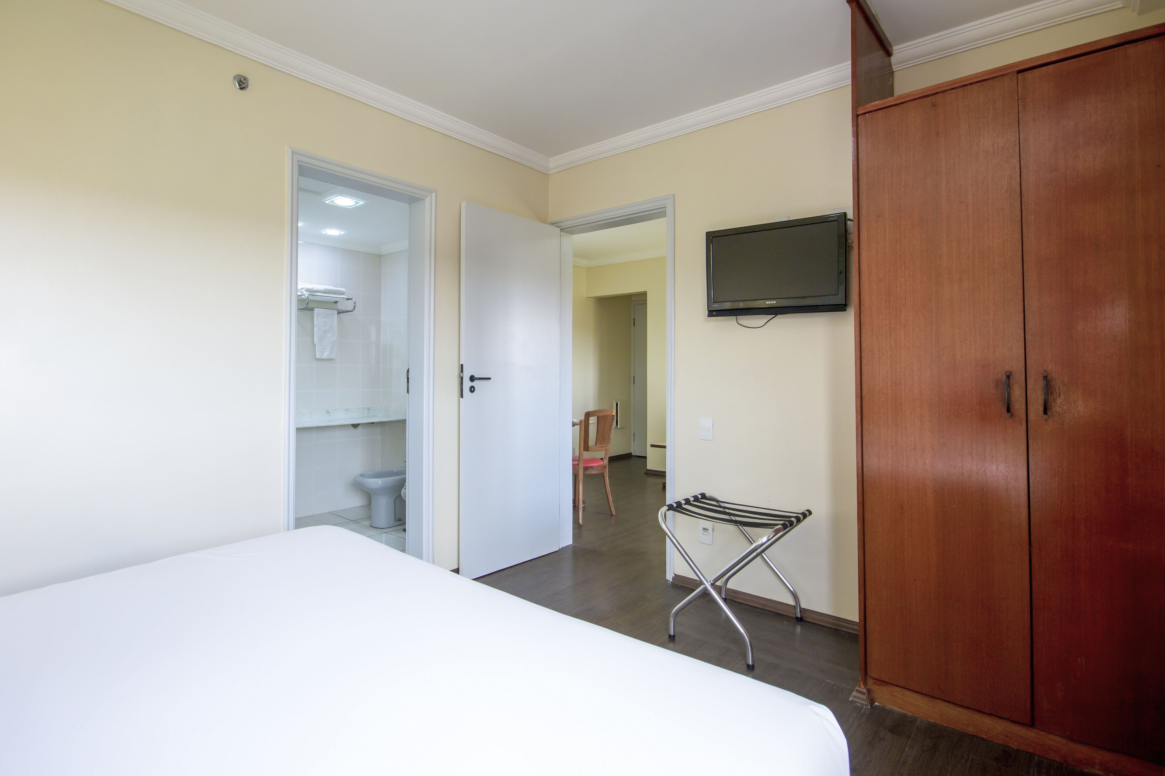 A bedroom with a bed, a wooden wardrobe, a television mounted on the wall, and a bathroom visible through an open door.