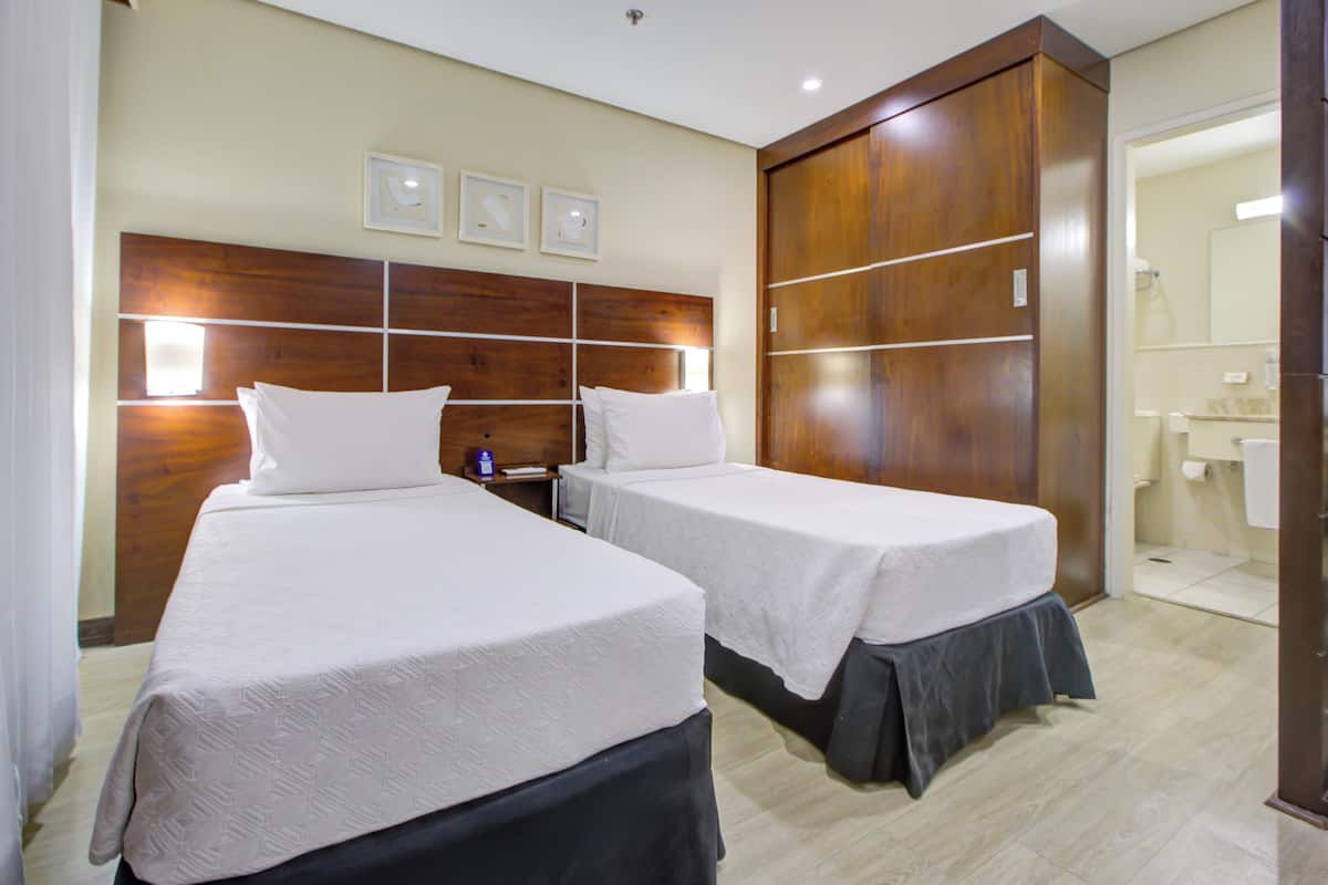 Habitación de hotel con dos camas, cabecera de madera, mesita de noche y baño visible a través de una puerta abierta.