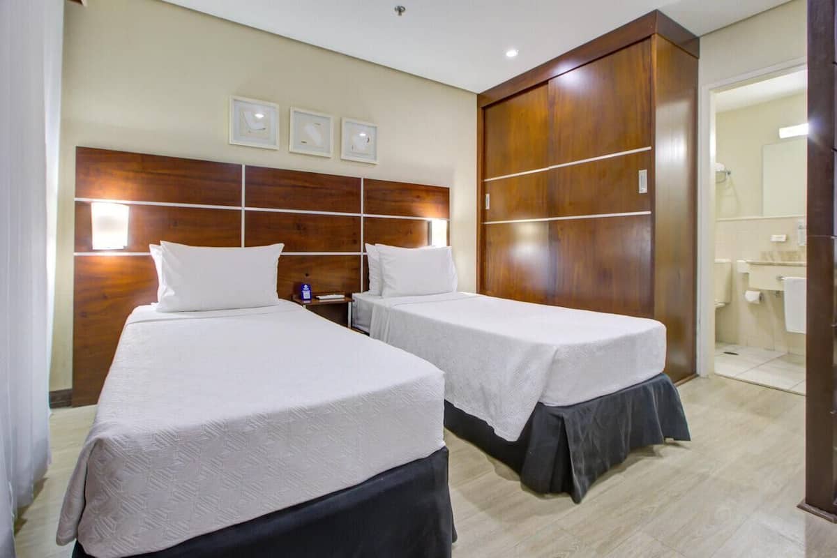 Habitación de hotel con dos camas, cabecero de madera, mesita de noche y baño visible a través de una puerta abierta.