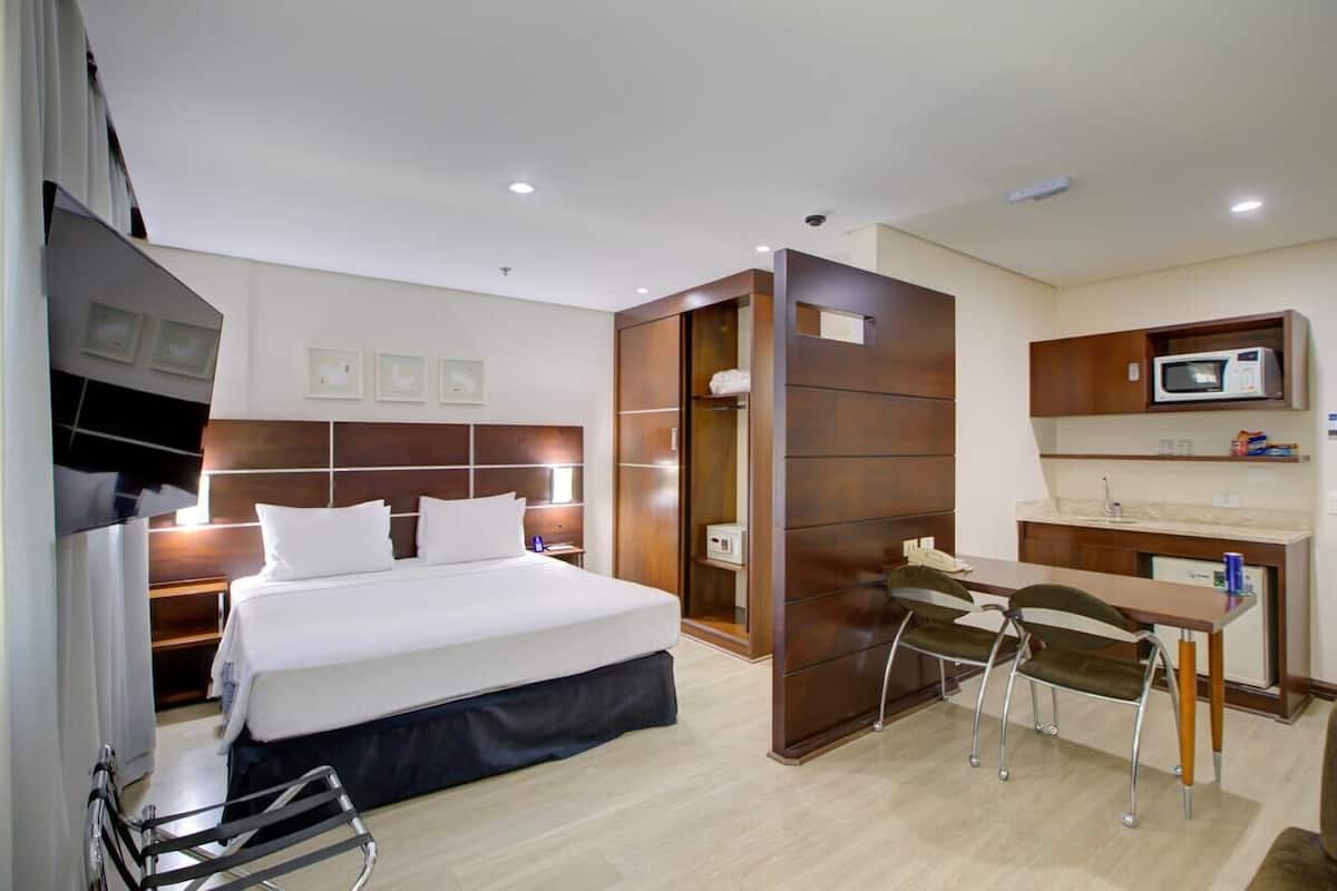 Habitación de hotel con cama, mesita de noche, escritorio con sillas, microondas y una pequeña kitchenette.