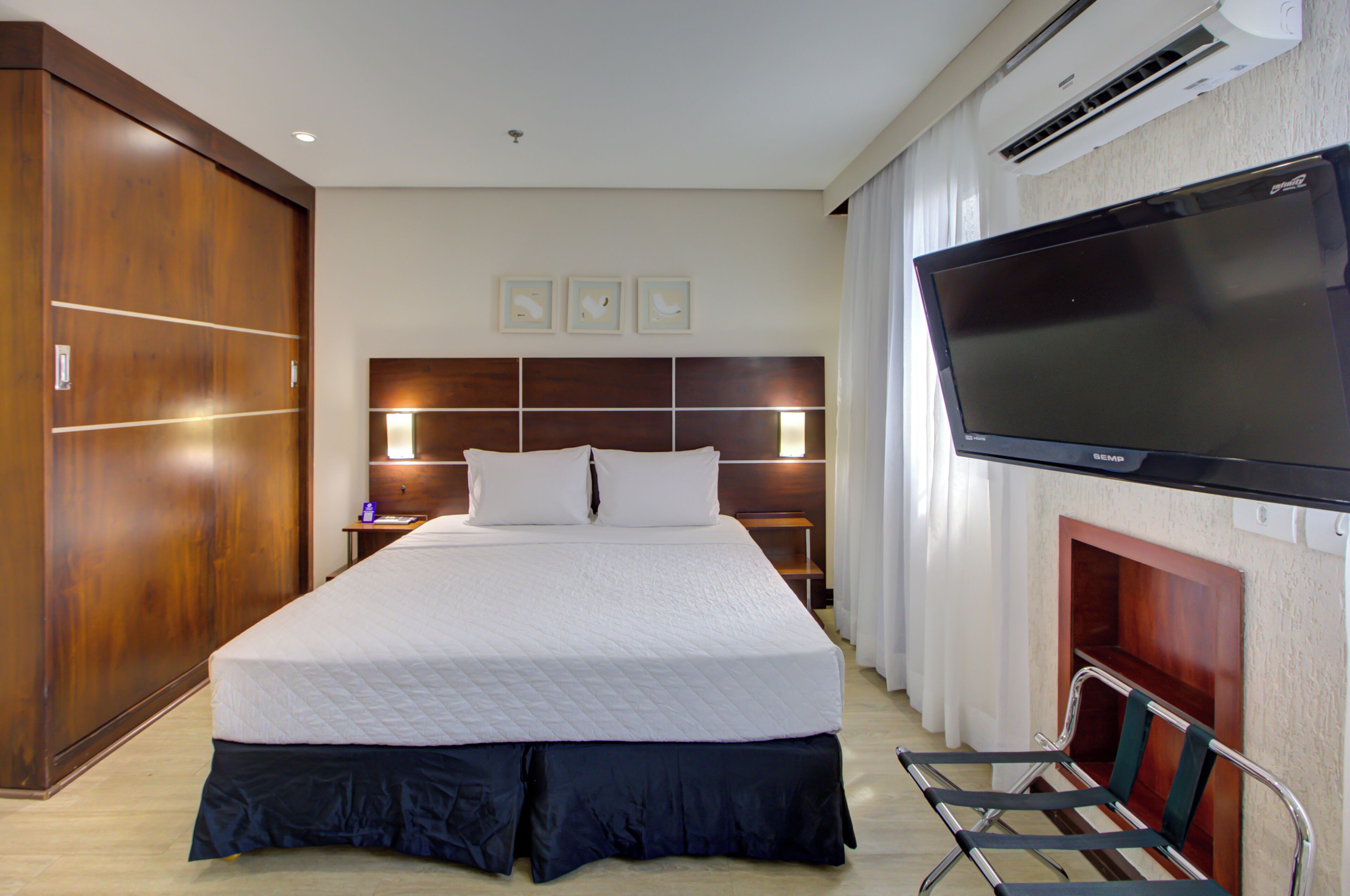 A hotel room with a large bed, a flat-screen TV mounted on the wall, a wooden wardrobe, and a chair.