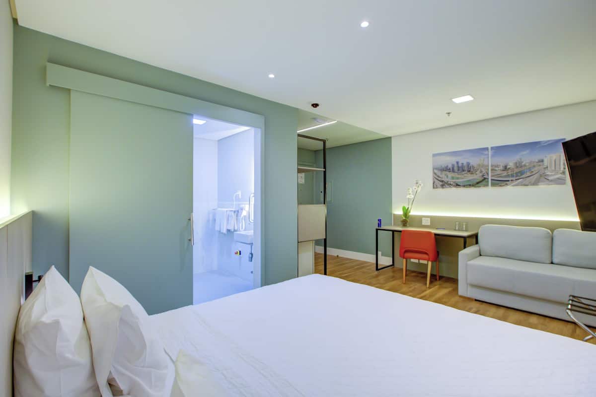 Una habitación de hotel con cama, escritorio, sillón, sofá y una pared decorada con una pintura de paisaje urbano.