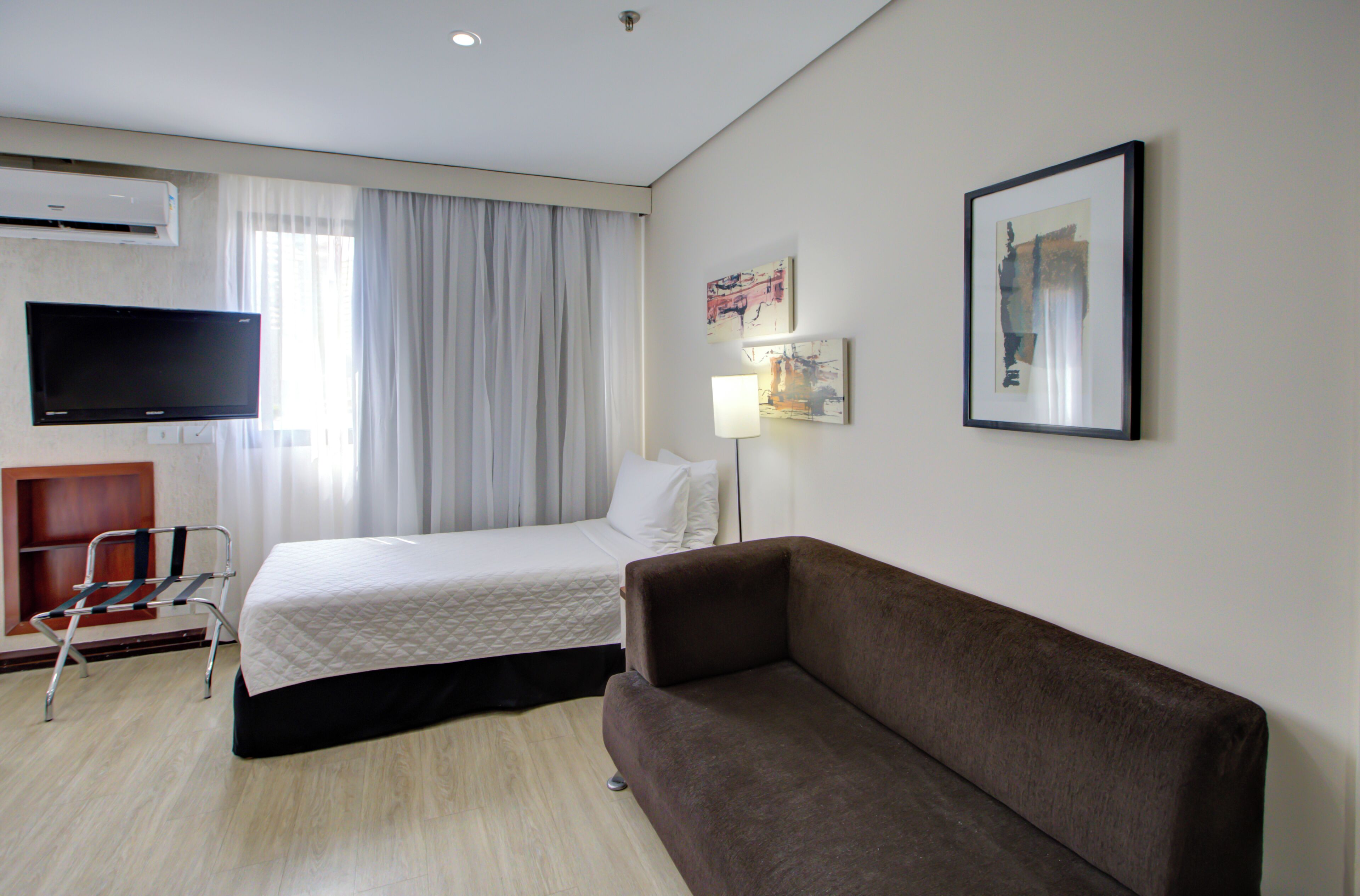 A hotel room with a bed, a sofa, a TV, a chair, and artwork on the walls.