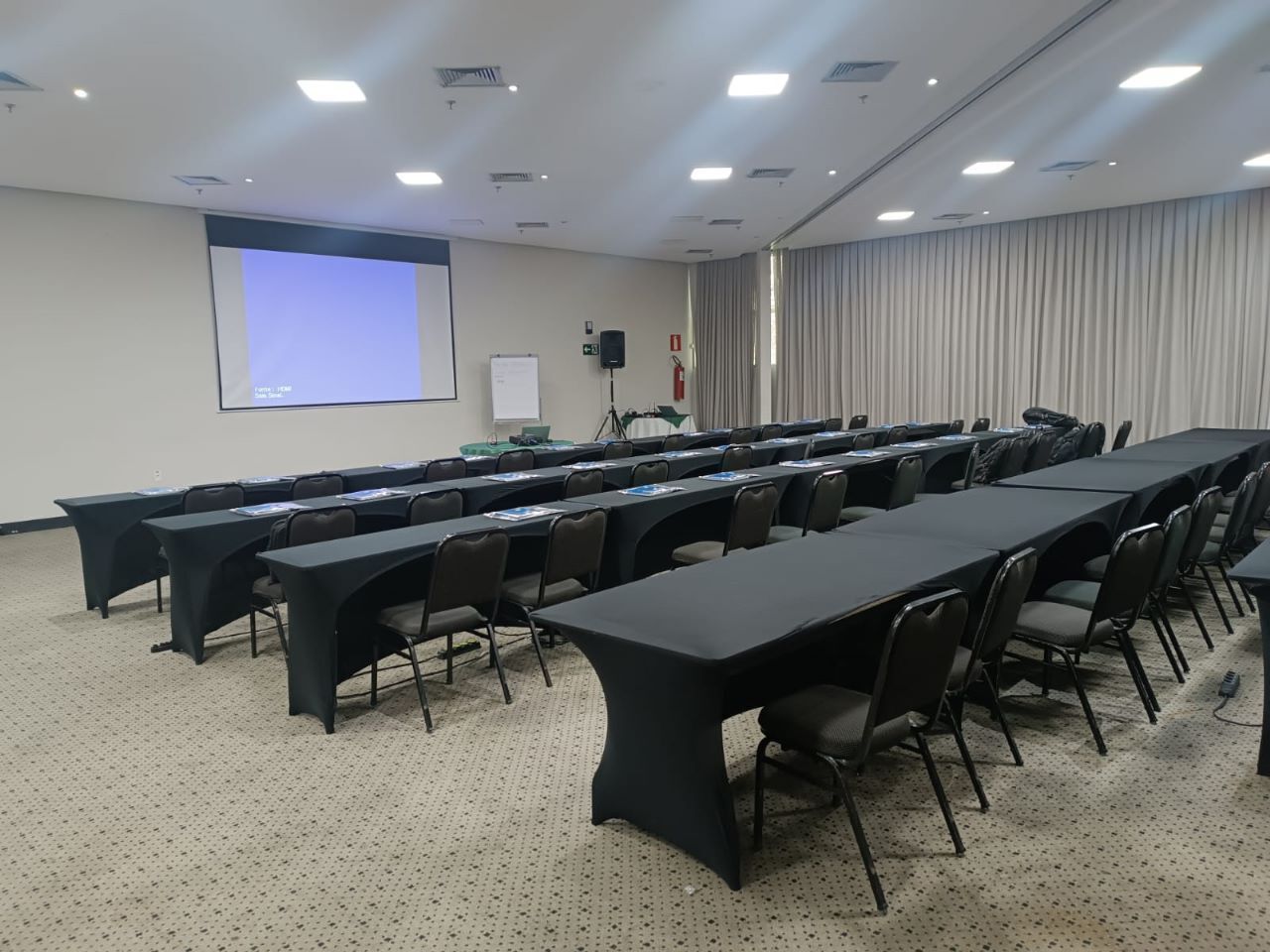 Un salón de conferencias con largas mesas, sillas, una pantalla de proyector y una pizarra.