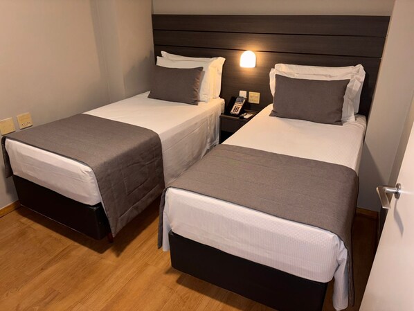 Dos camas individuales con ropa de cama gris, una mesita de noche con un teléfono y una lámpara, y suelo de madera.
