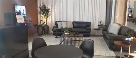 Um hall de hotel moderno com balcão de recepção, sofás de couro preto, uma mesa redonda e plantas em vasos.