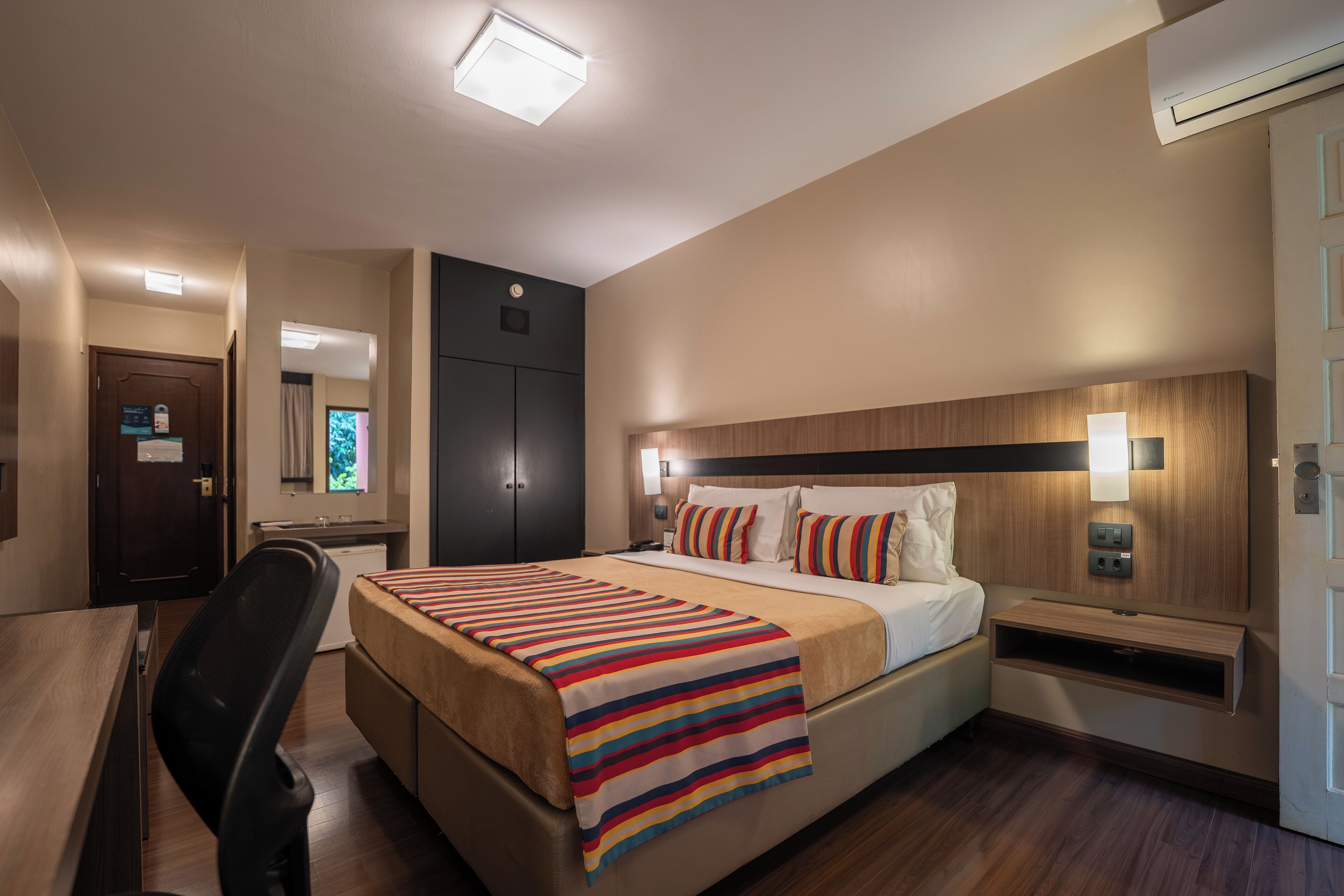 Una habitación de hotel con cama, mesita de noche, escritorio, silla y ventana con cortinas.