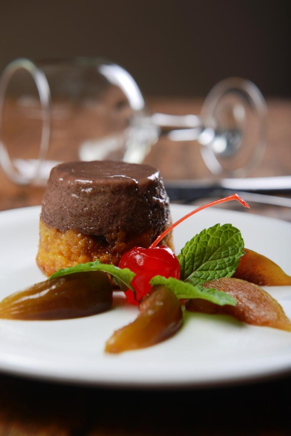 A dessert with a chocolate topping, garnished with mint leaves and a cherry, on a white plate.