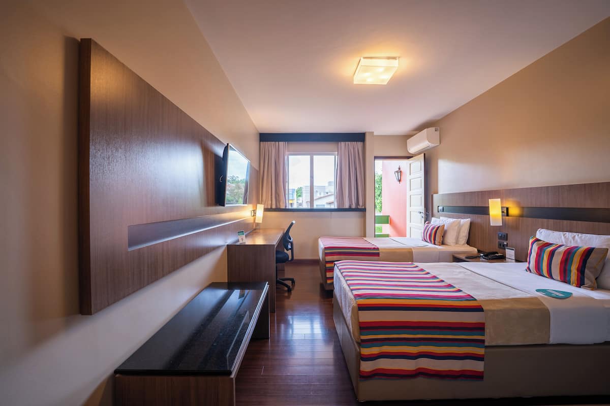 Habitación de hotel con escritorio, dos camas, un banco, televisor y ventana con cortinas.