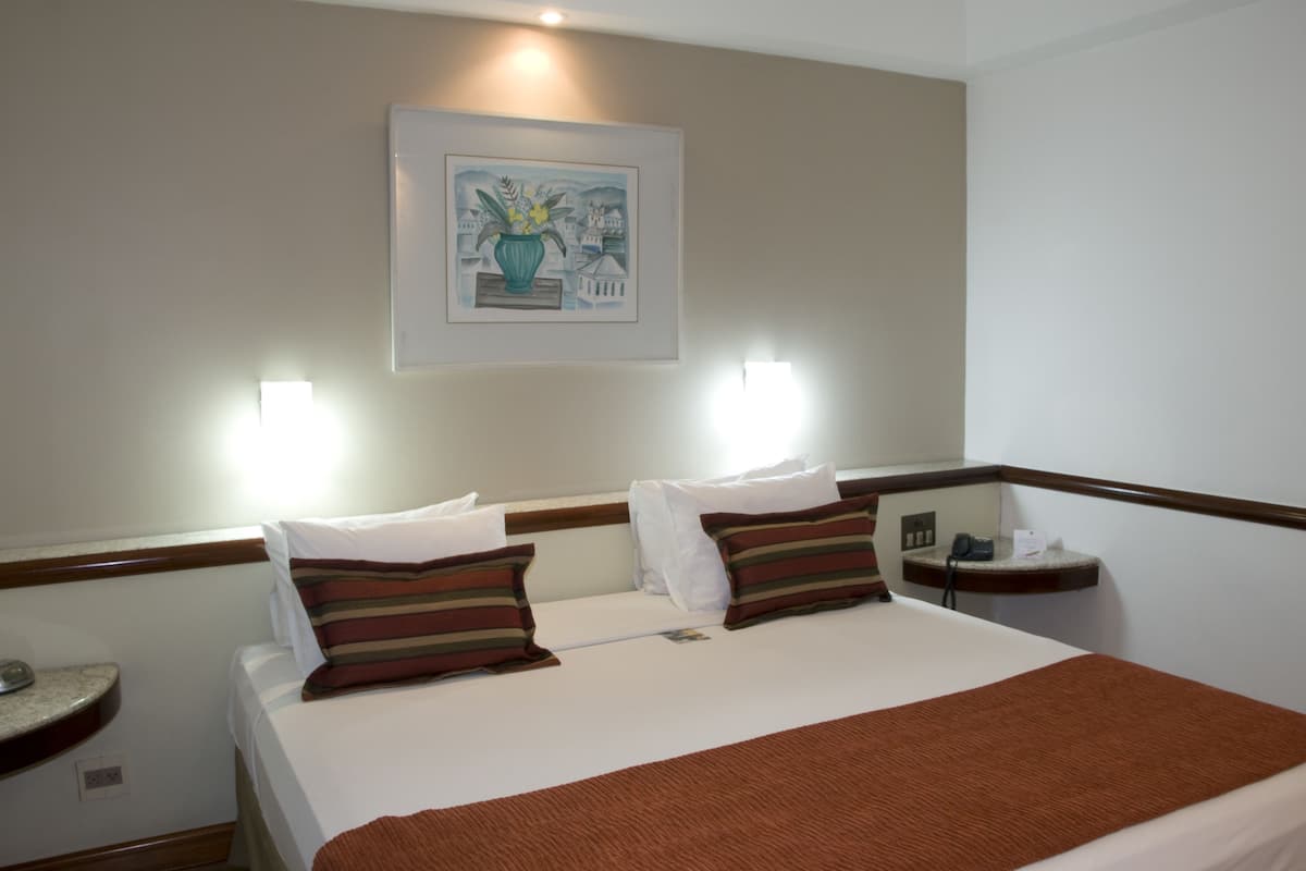 Habitación de hotel con una cama, dos mesitas de noche, una lámpara de pared y un cuadro enmarcado.