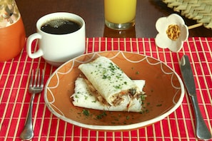 A plate with a rolled dish, a cup of coffee, and a glass of orange juice on a red and white checkered tablecloth.