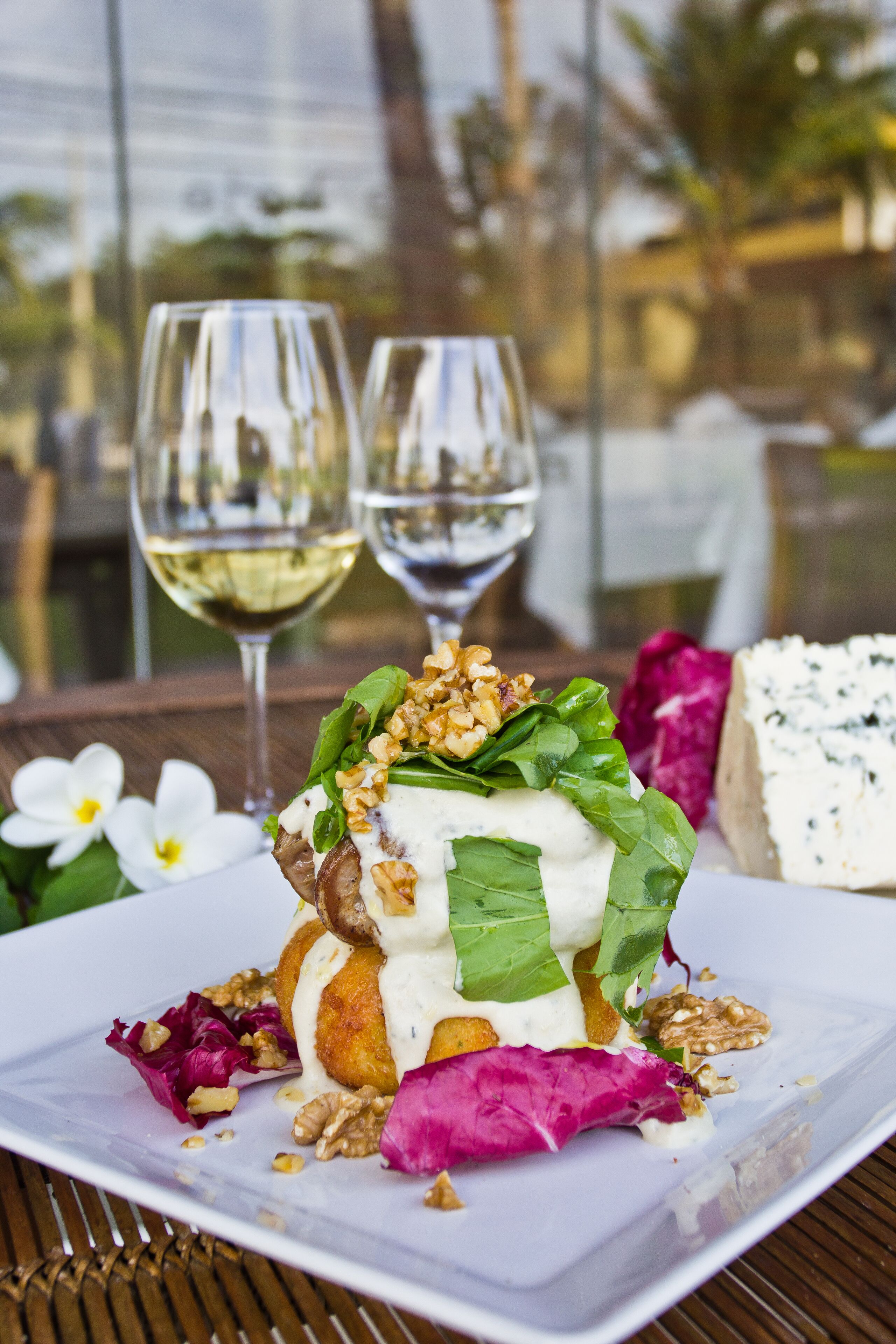 A plated dish with greens, nuts, and a creamy topping, accompanied by a glass of white wine, set on a table with a view of palm trees and buildings.
