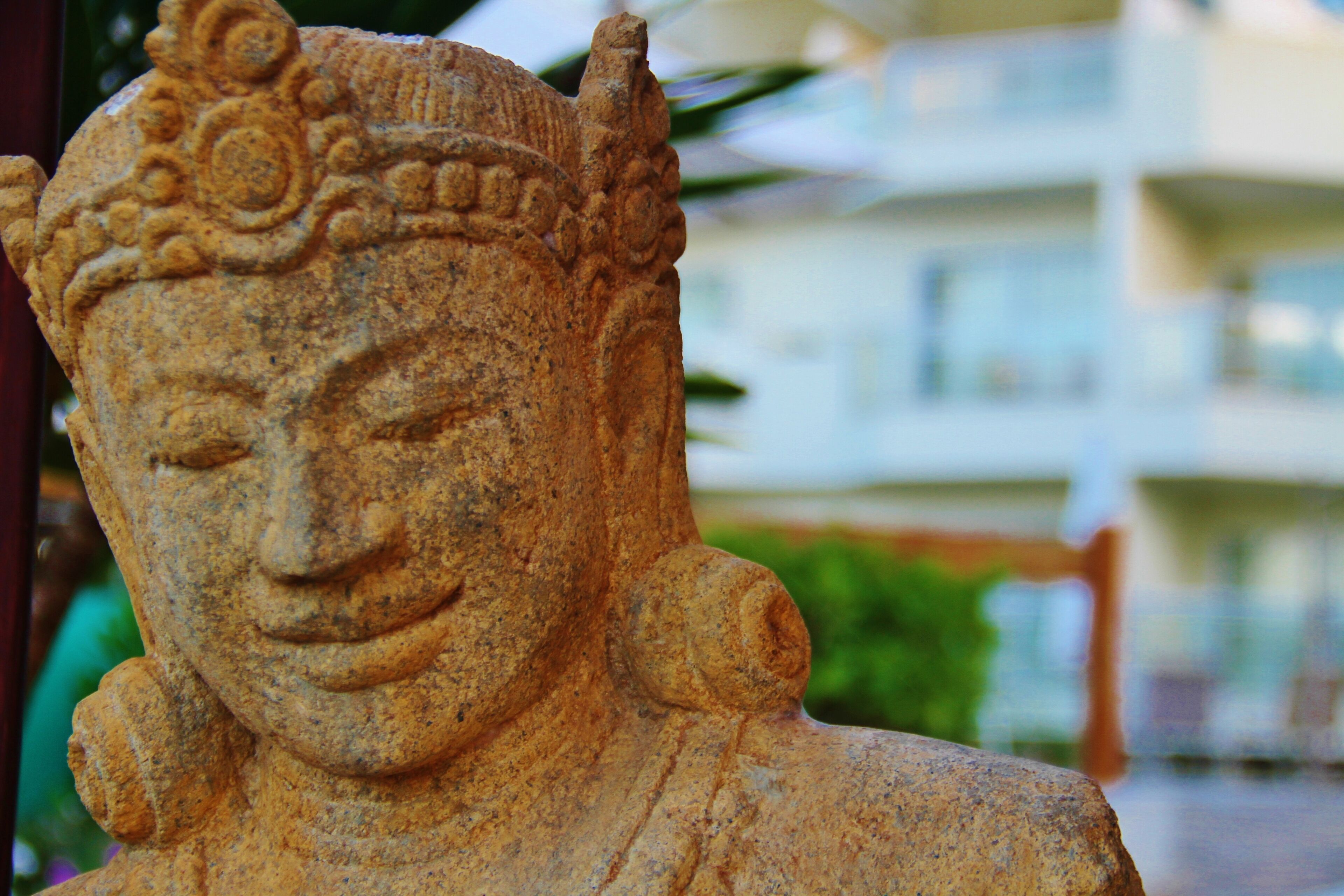 A stone statue with intricate carvings, set against a blurred background of a building and greenery.