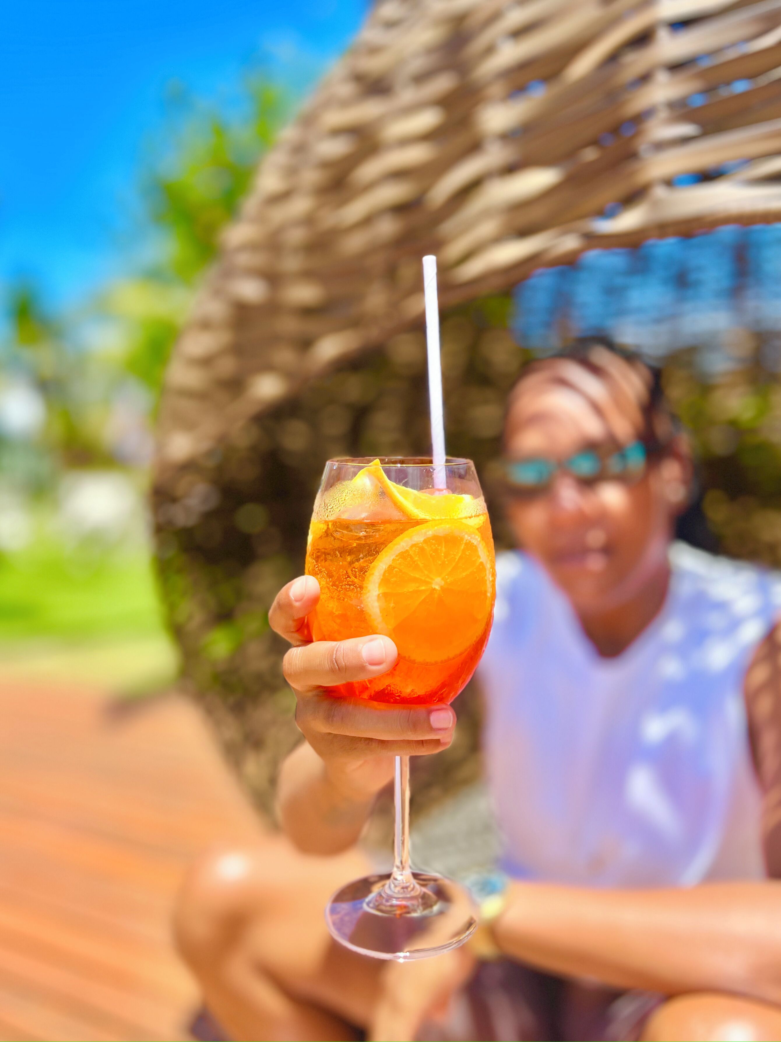 A person holding a glass with a straw, containing a beverage with an orange slice, outdoors.
