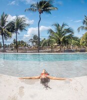 Vista de la piscina con agua cristalina, palmeras y una persona recostada en el borde.