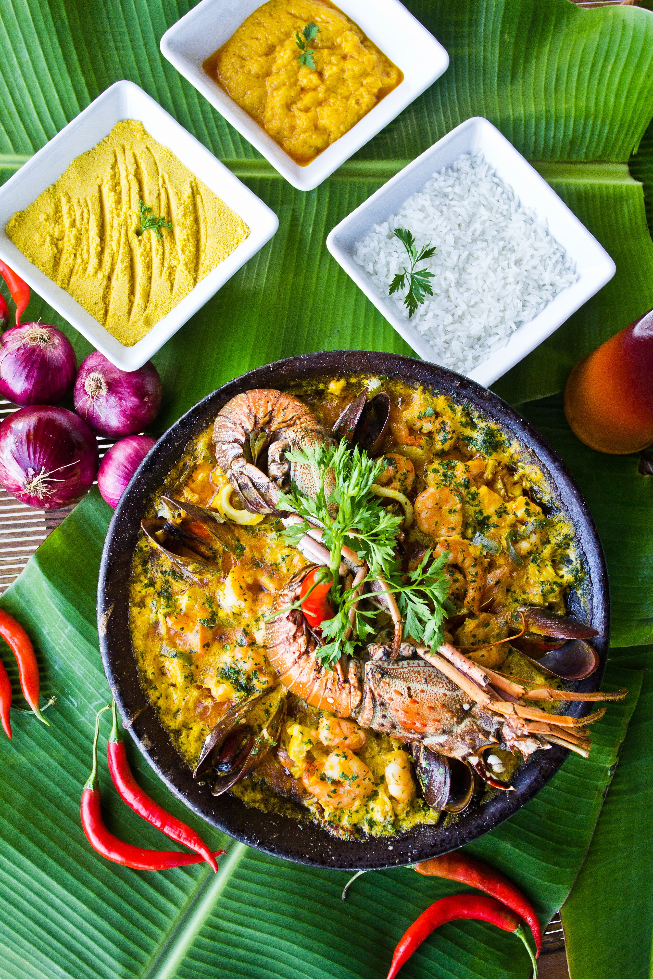 A seafood dish with mussels and shrimp, accompanied by bowls of rice and a yellow sauce, set on a banana leaf.