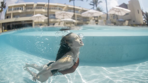 An underwater view of a swimming pool with clear water, a person swimming, and a resort building with palm trees and umbrellas in the background. - Casa Di Vina Boutique Hotel (Salvador)