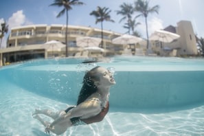 Vista subacuática de una piscina con agua cristalina, una persona nadando y un edificio de resort con palmeras y sombrillas al fondo.