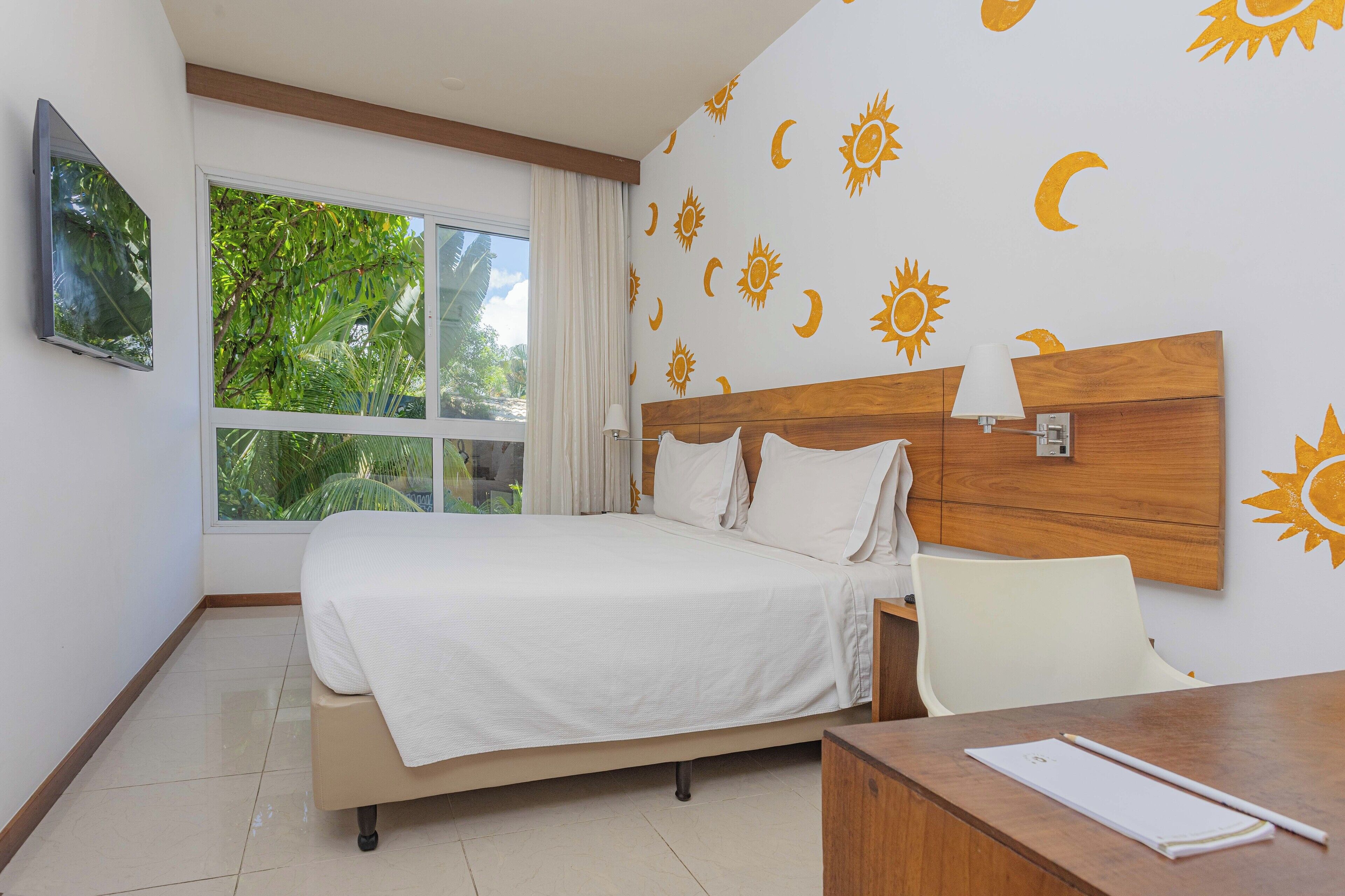 A hotel room with a bed, a desk, and a window with a view of greenery.